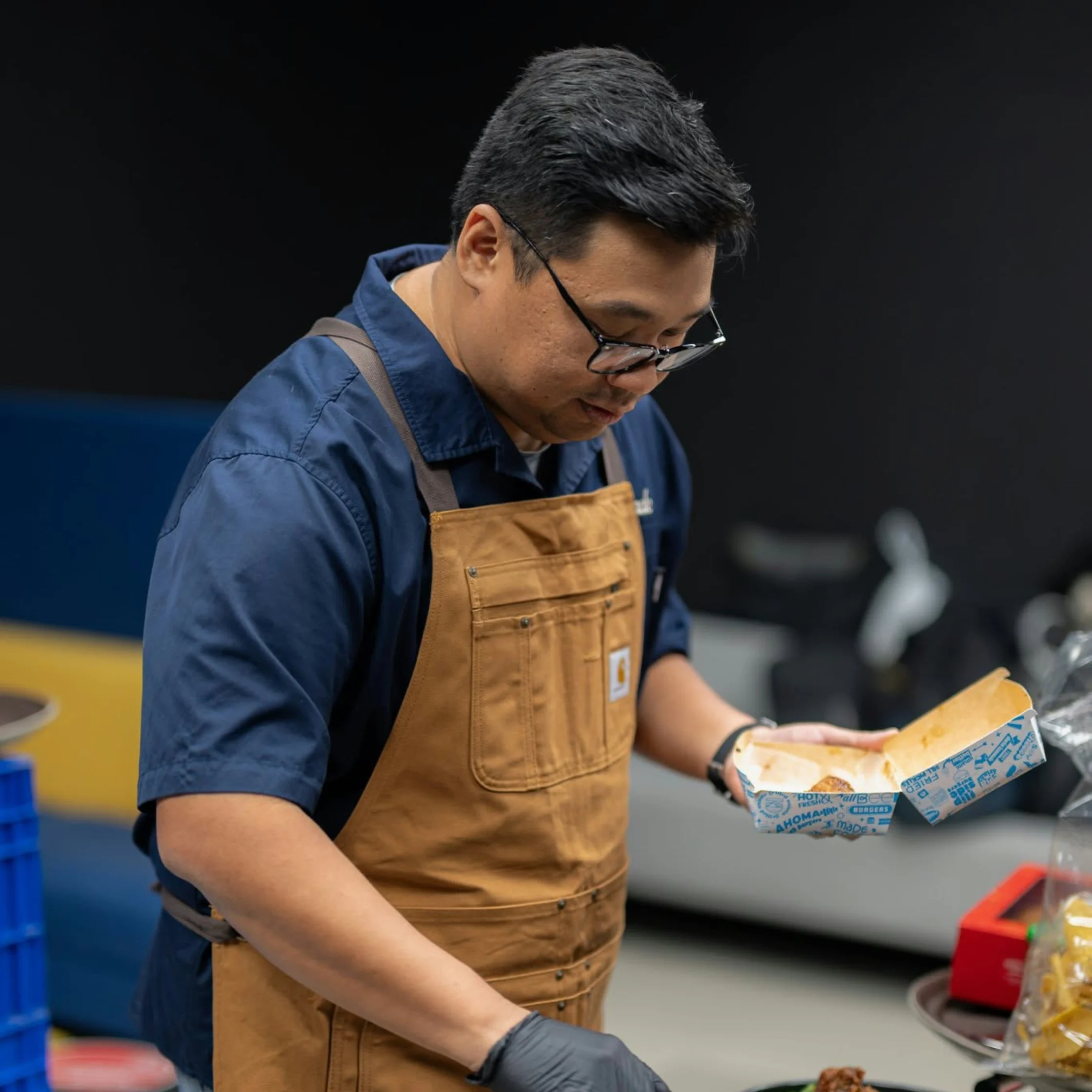 A focused chef in a blue shirt and brown apron examines a wrapped ingredient at a kitchen counter, conveying calm concentration in a working kitchen.