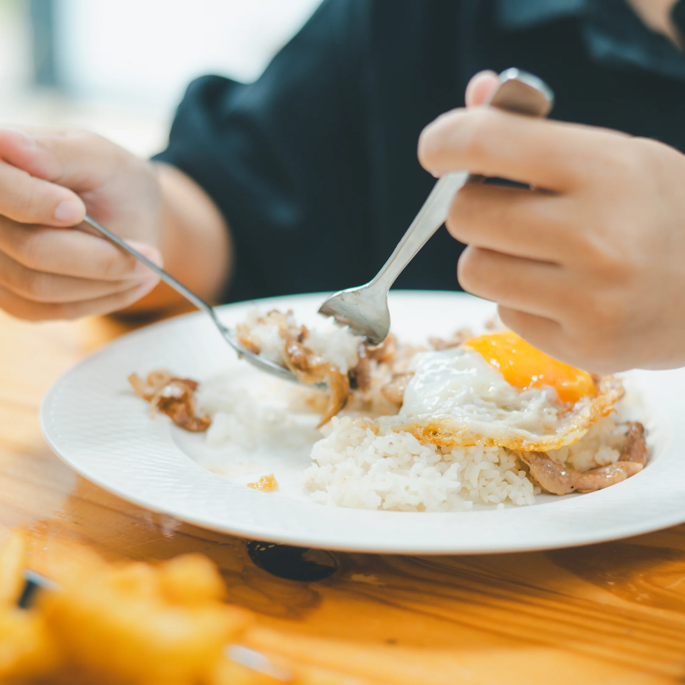 A person in dark clothing uses a fork and spoon to eat rice topped with a sunny-side-up egg and meat, in a relaxed dining setting.