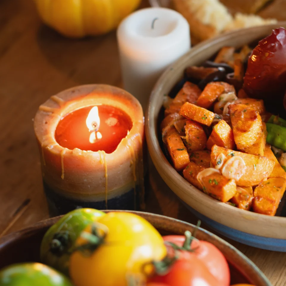 A glowing candle beside bowls of roasted orange vegetables, tomatoes, and peppers, with a pumpkin in the background, creating a warm, autumnal mood.