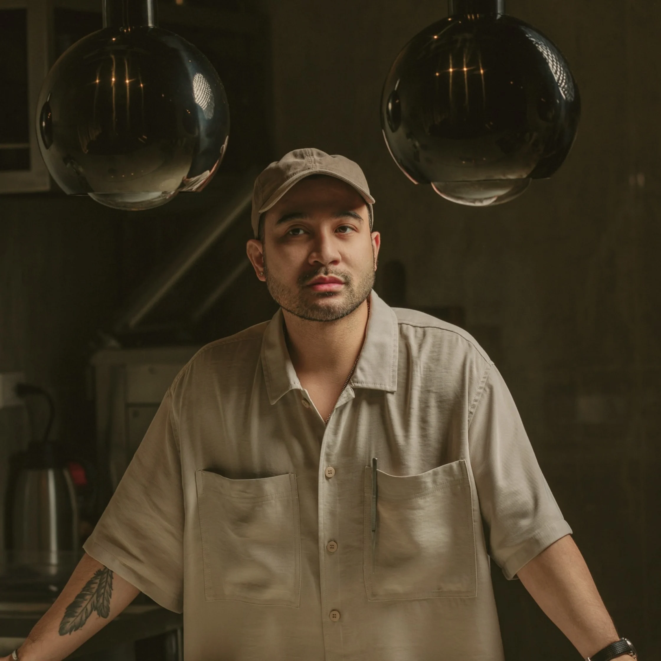 A thoughtful man in a beige cap and work shirt leans on a counter beneath two glossy pendant lights in a dim industrial kitchen.