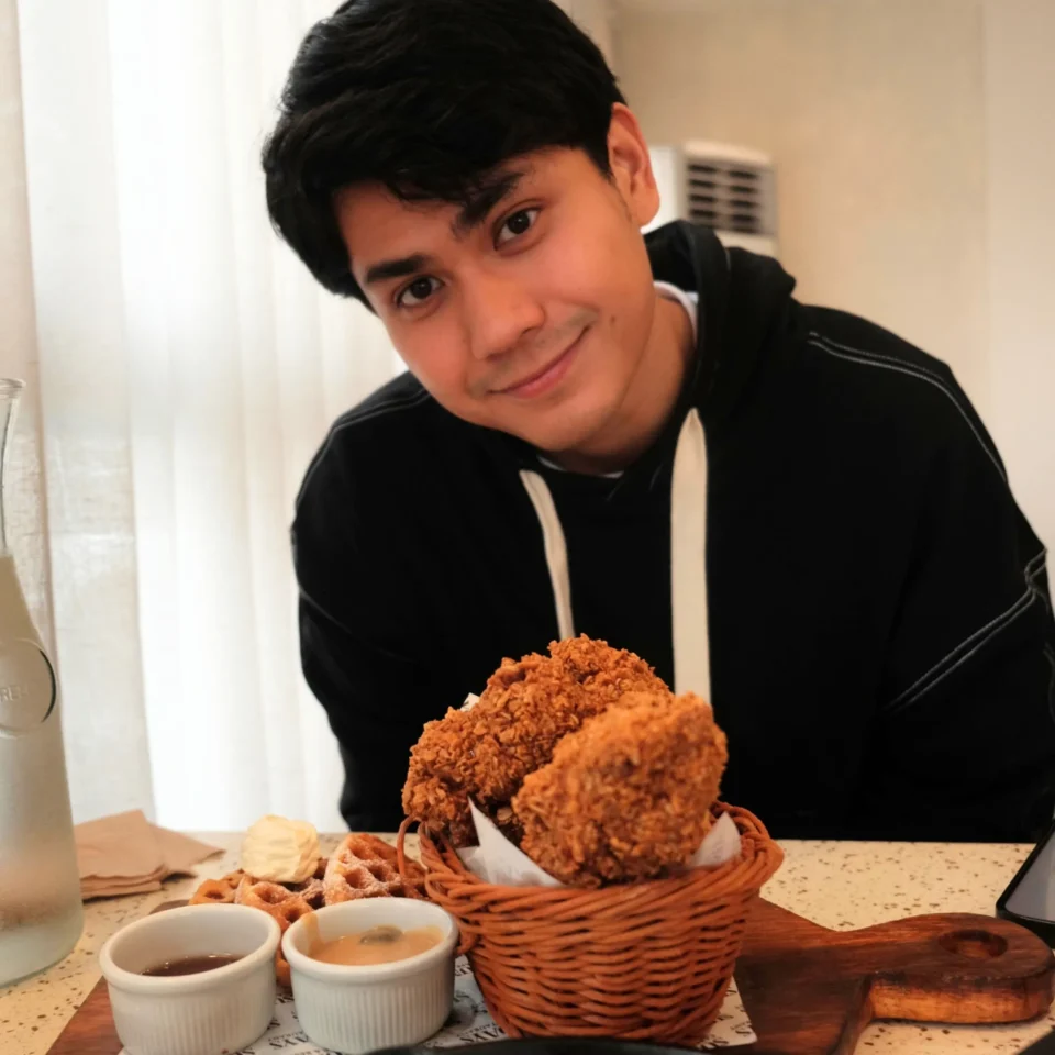 A young man in a black hoodie smiles at the camera behind a table with a basket of fried snacks, dipping sauces, and a wooden serving board.