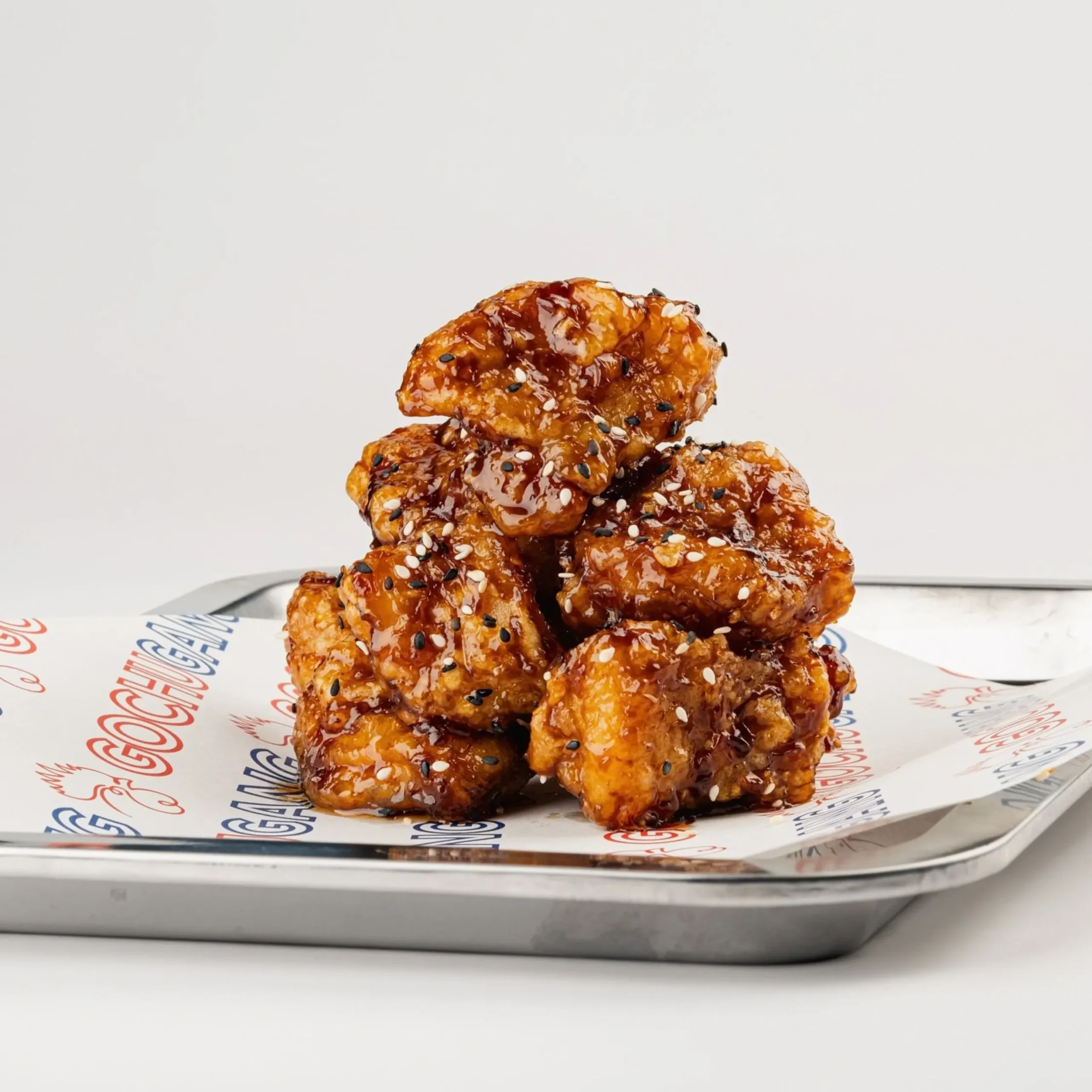 A stack of crispy, glazed chicken wings coated in glossy sauce and sesame seeds sits on branded paper over a metal tray, looking savory and appetizing.