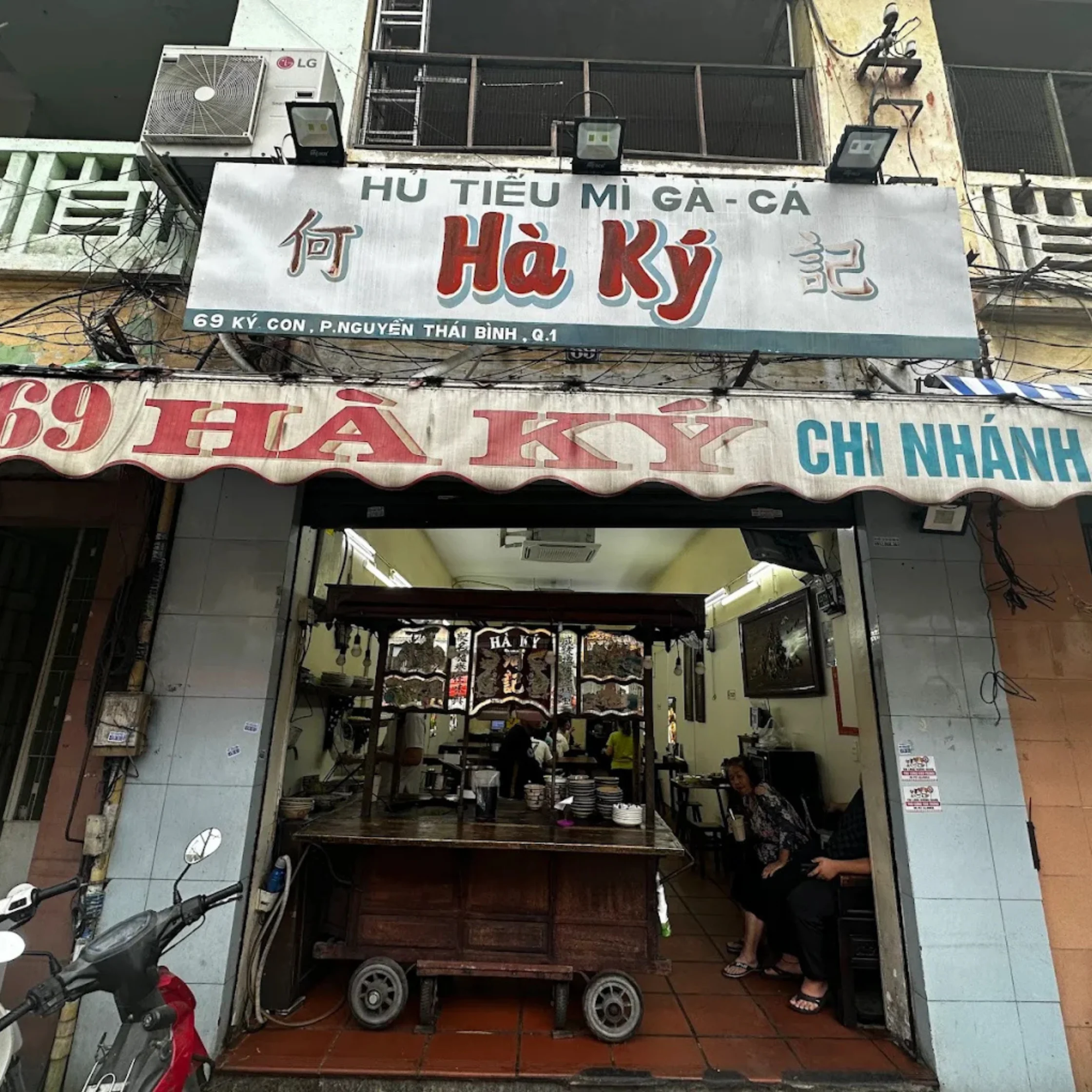 Vietnamese noodle shop Hảo Ký with weathered signs and open entrance; a food cart and parked scooters evoke a modest, bustling street-side eatery.