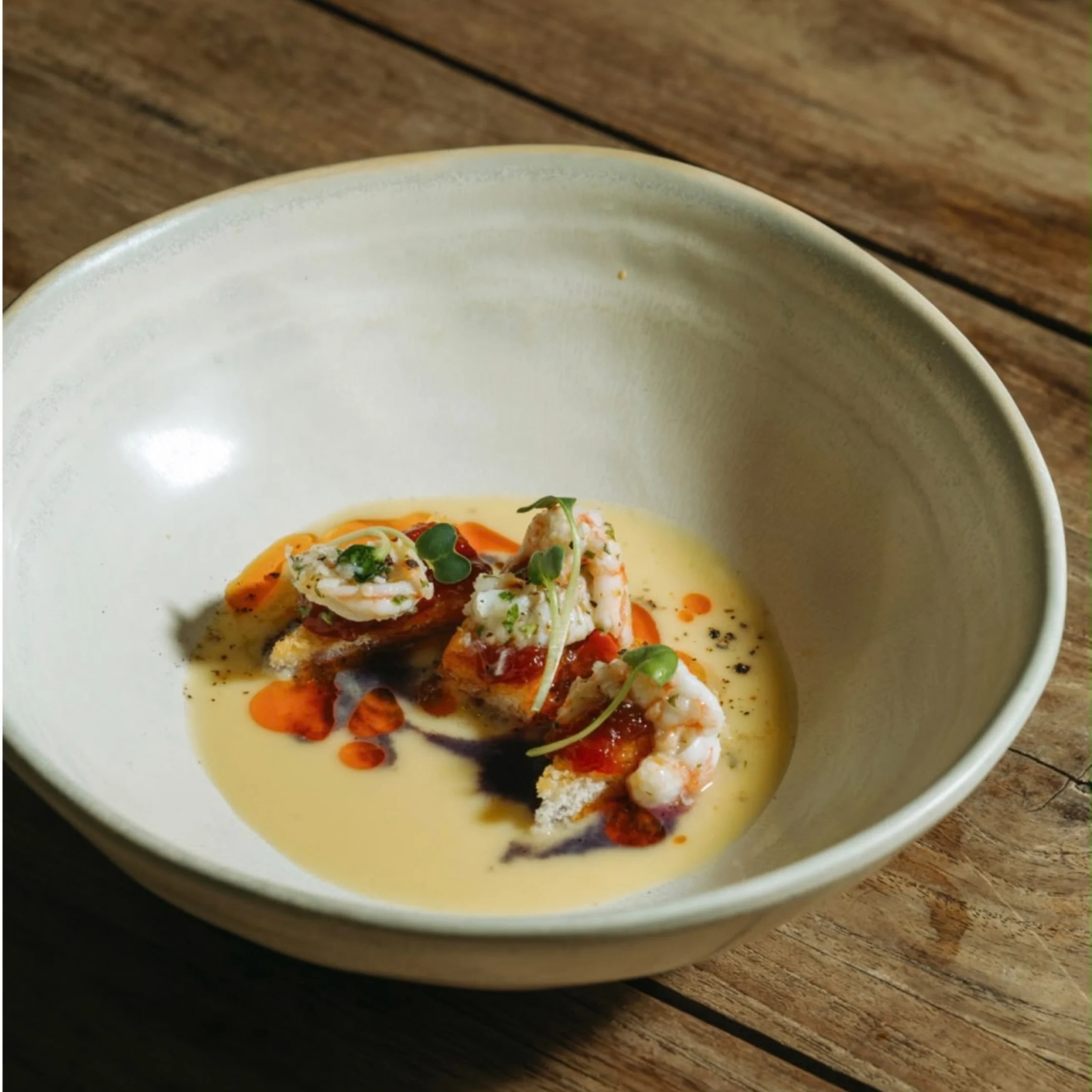 A shallow ceramic bowl holds shrimp in creamy yellow sauce with red oil, herbs, and toasted crumbs on a rustic wooden table; elegant, intimate plating.