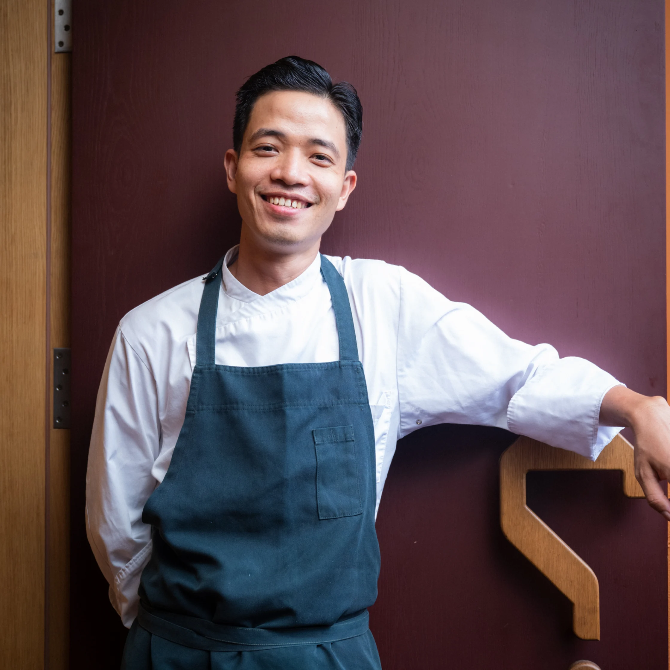 A smiling chef in a white jacket and dark blue apron leans against a wooden counter before a burgundy wall, conveying a warm, welcoming mood.