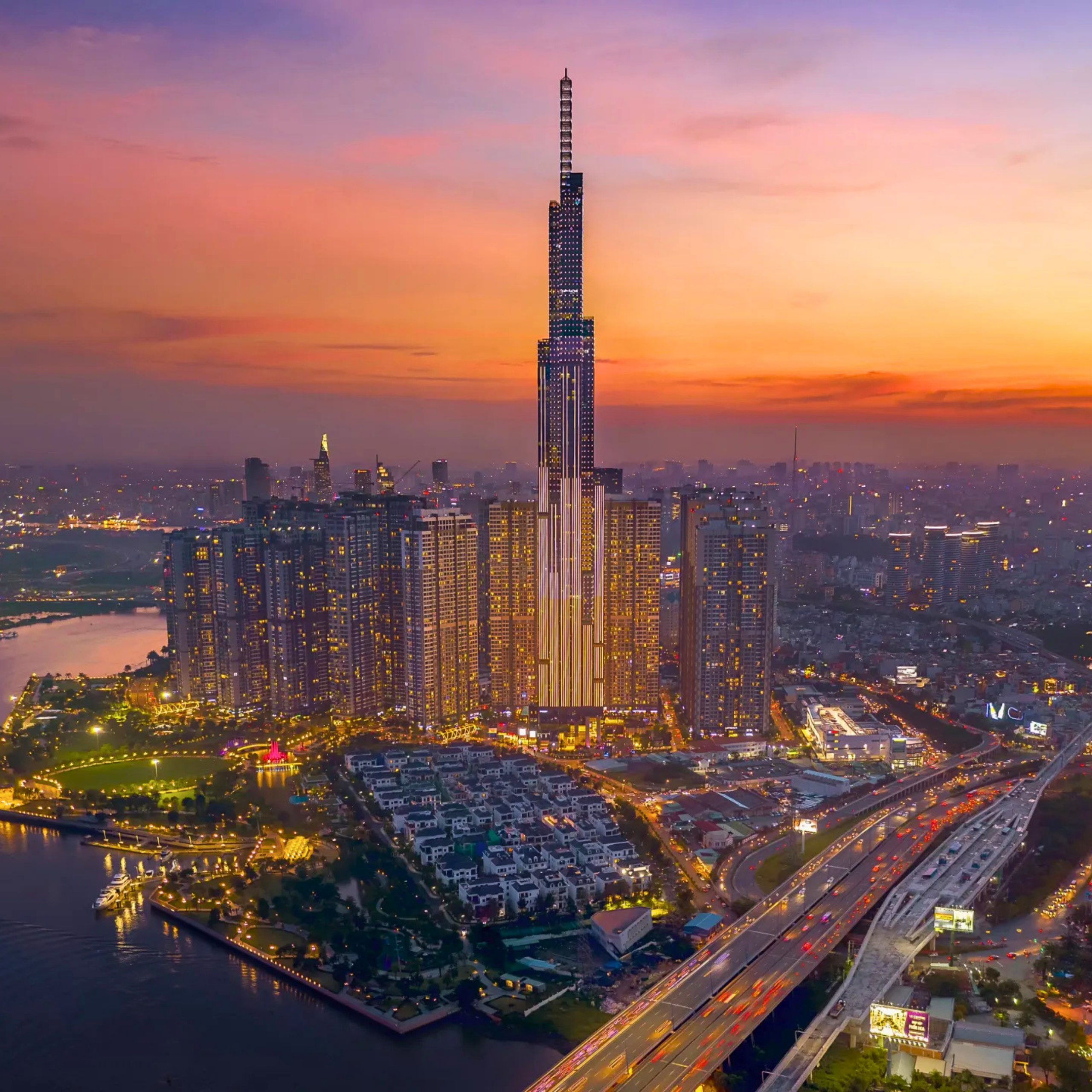 Aerial view of Ho Chi Minh City at sunset, with the tall skyscraper rising above dense skyscrapers, highways, and waterways beneath an orange-pink sky.