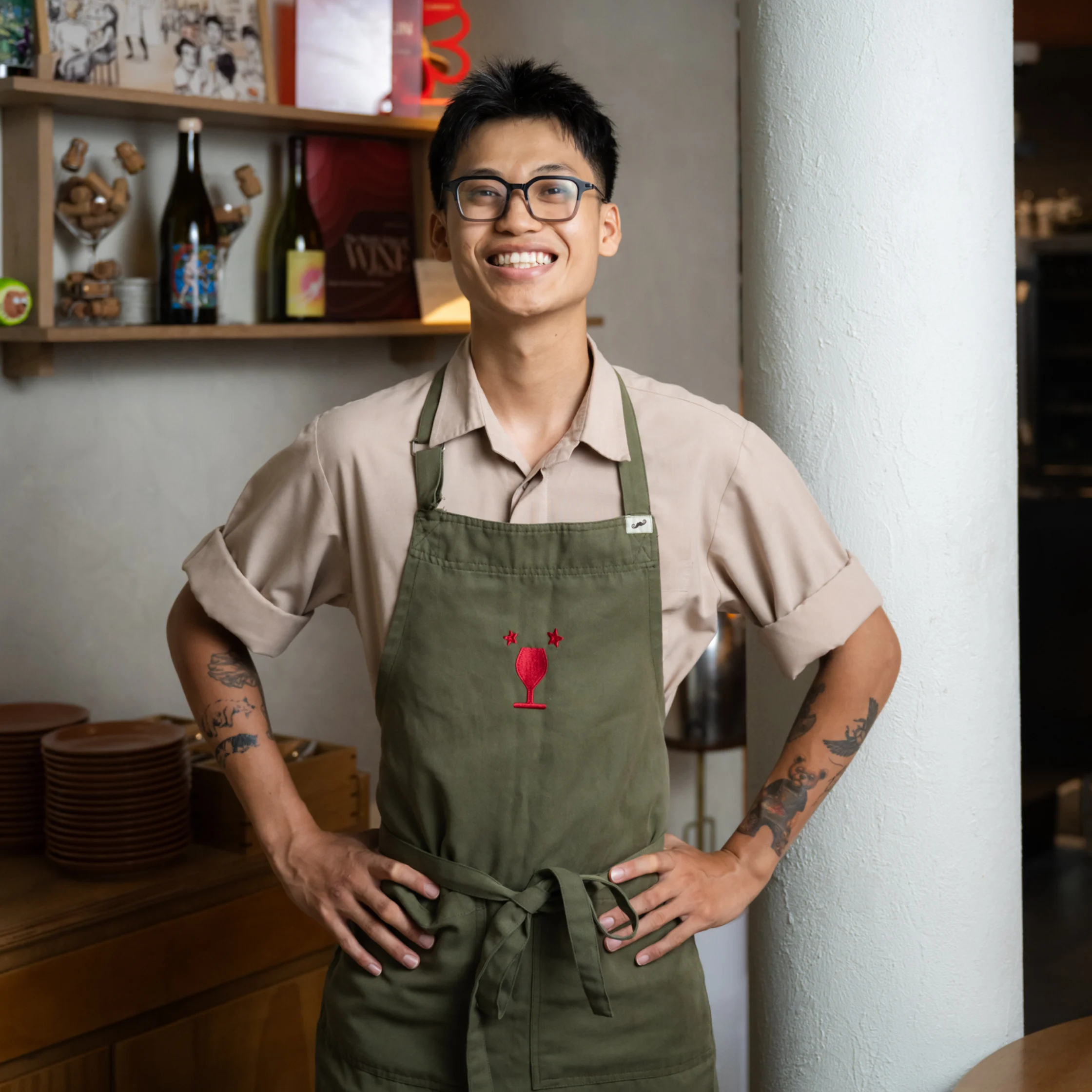 A smiling restaurant worker in glasses and a green apron stands with hands on hips in a cozy café, conveying warmth and confidence.
