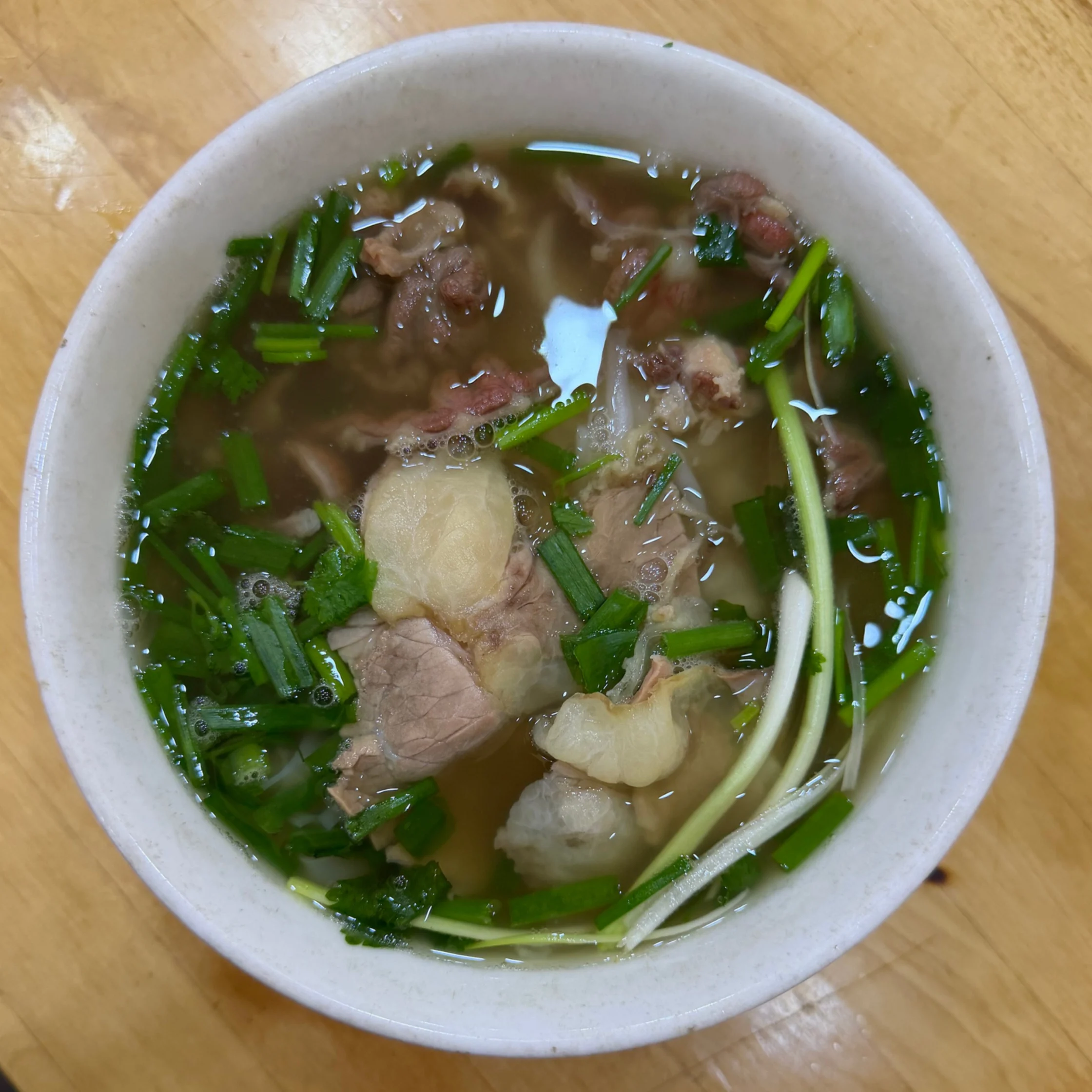 Overhead view of a white bowl of clear beef soup with meat, pale bone, chopped green herbs, and scallions on a wooden table; warm, comforting meal.