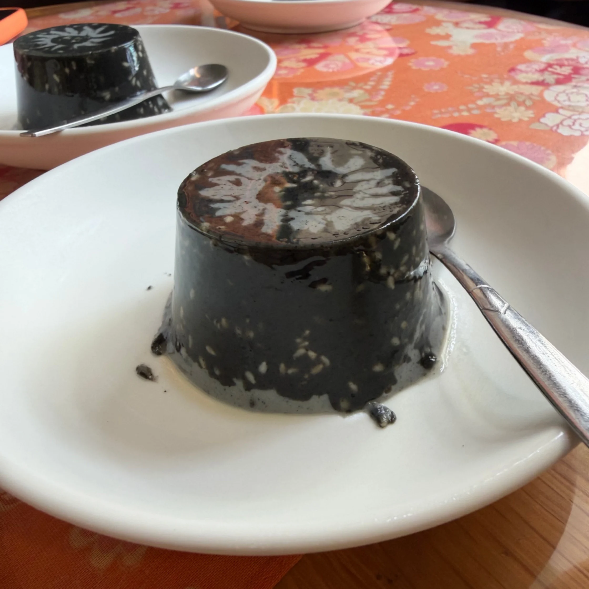 A glossy dark sesame pudding with pale flecks sits on a white plate beside a spoon, on a floral tablecloth; a casual, inviting dessert scene.