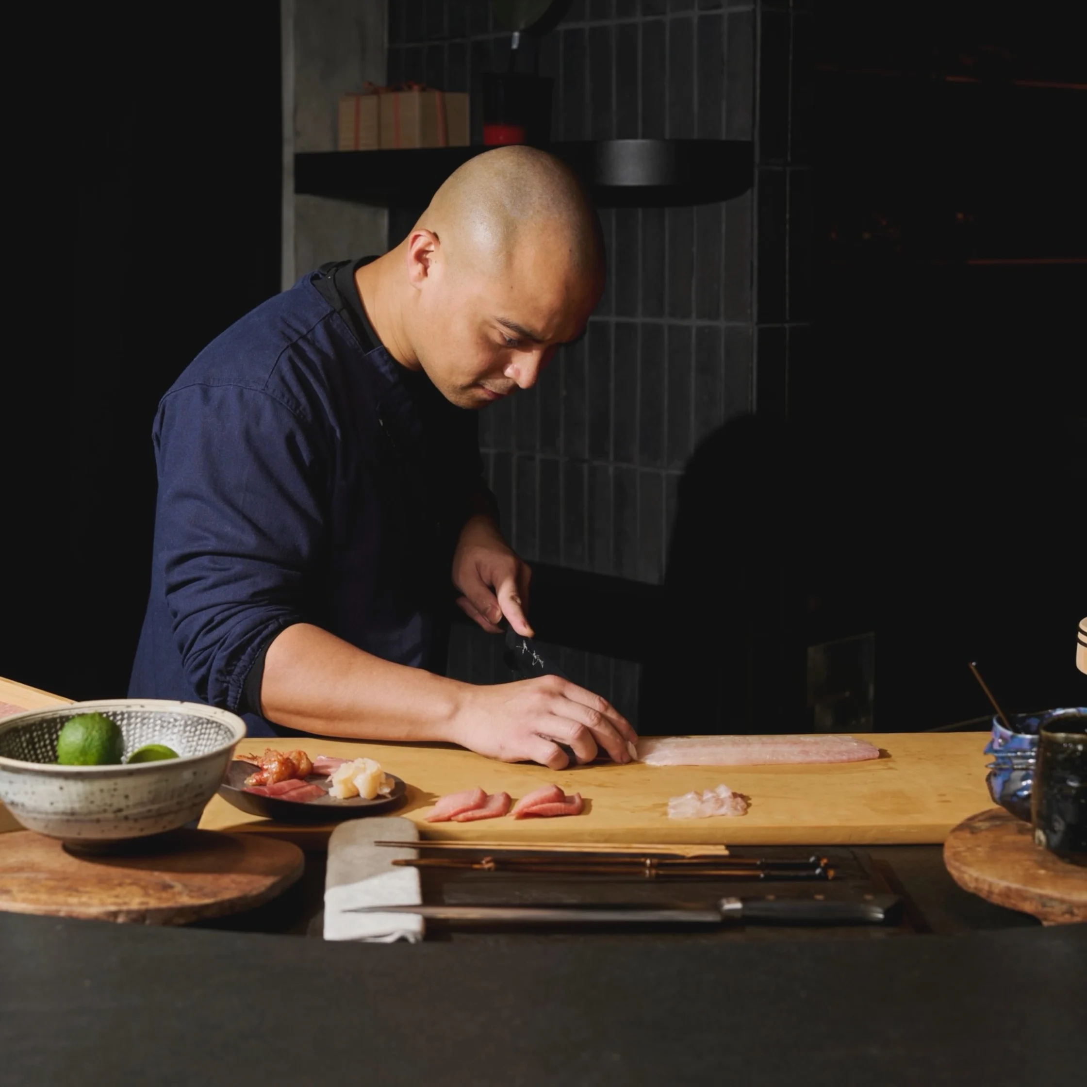 A focused chef in a dark blue shirt slices meat on a wooden cutting board in a dim kitchen, with bowls and ingredients nearby; calm, intimate mood.