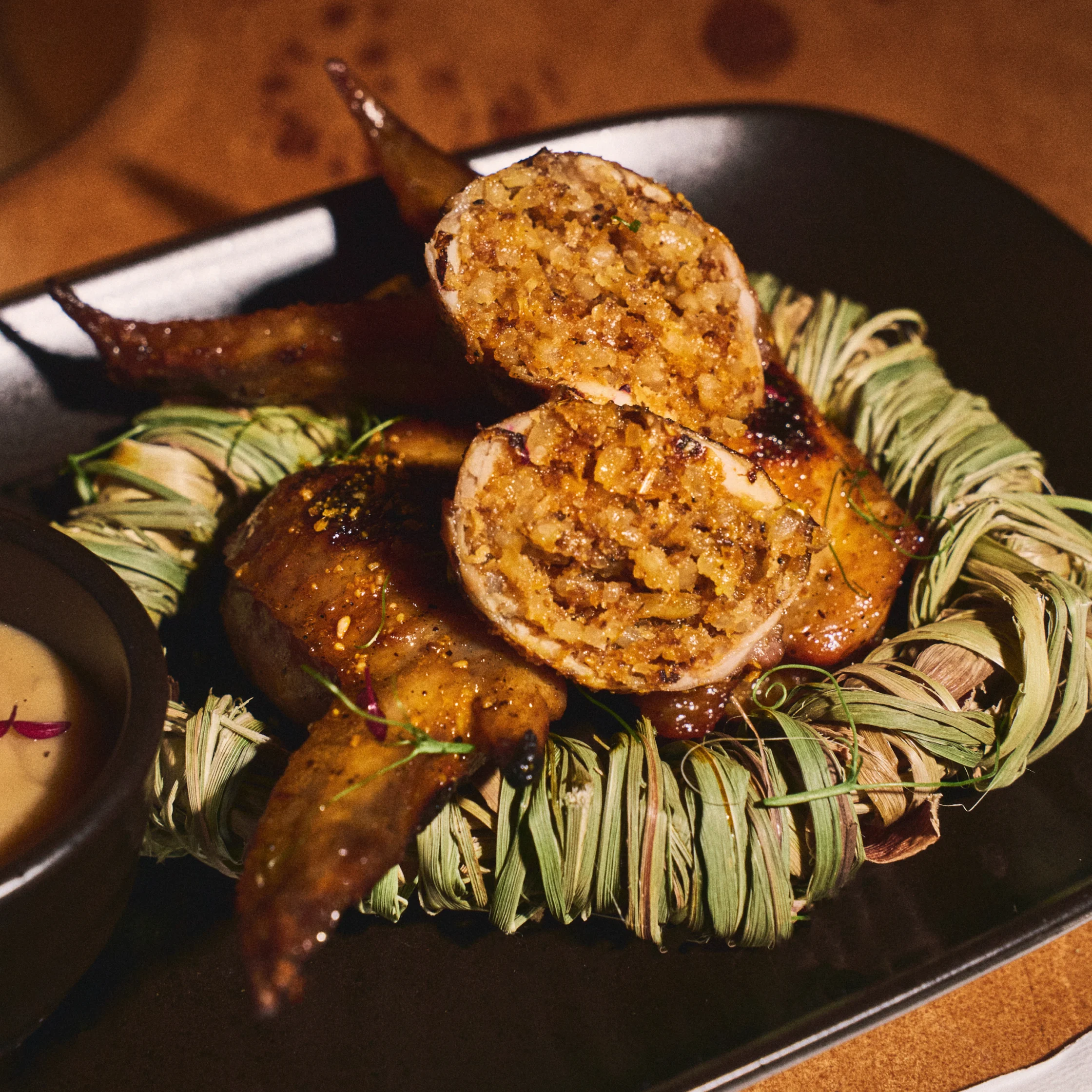 A richly browned, stuffed poultry dish is sliced open on a black plate, revealing savory filling, surrounded by herbs and served with dipping sauce.