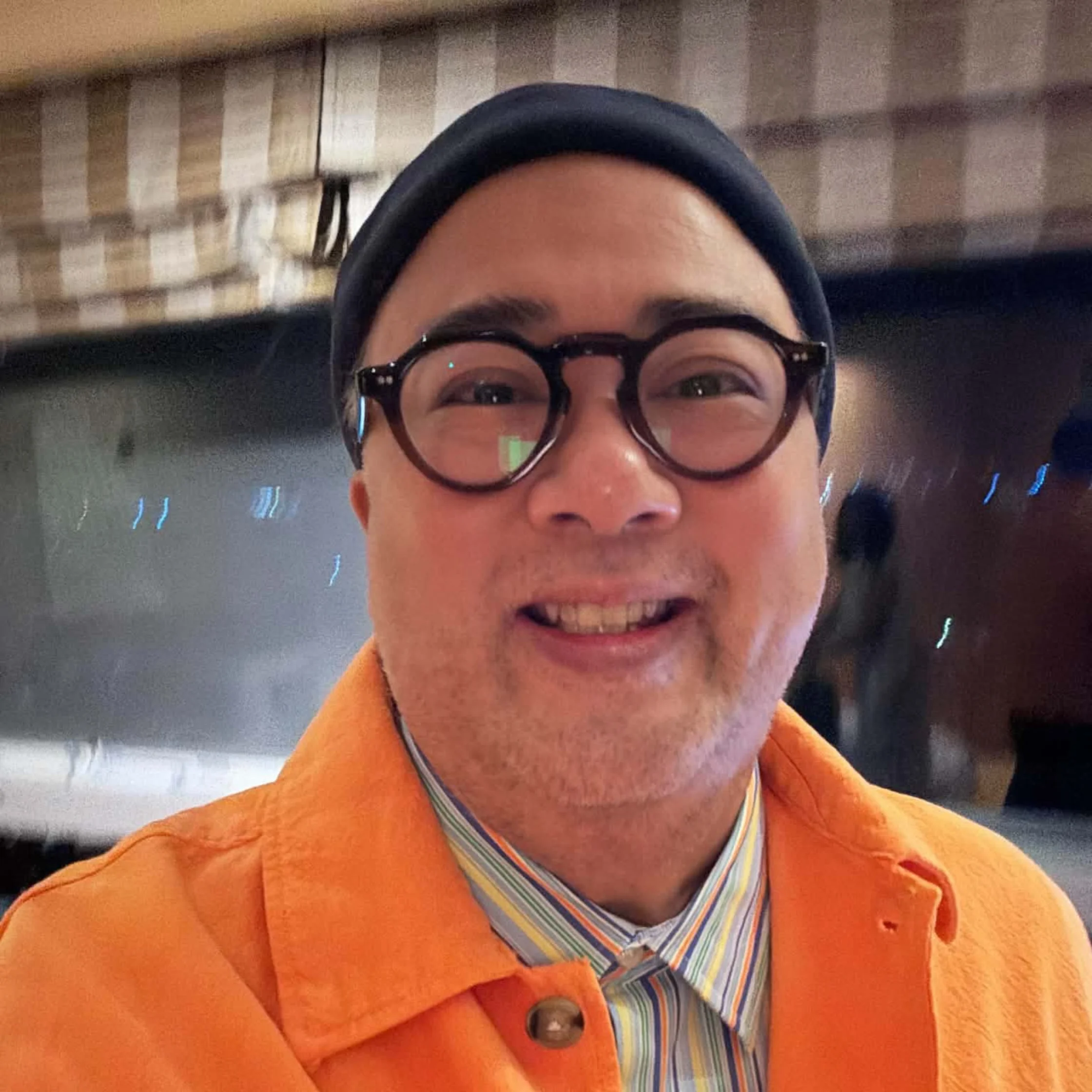 A smiling person in round black glasses and a black beanie wears a bright orange jacket over a striped shirt, posing indoors in warm, casual surroundings.