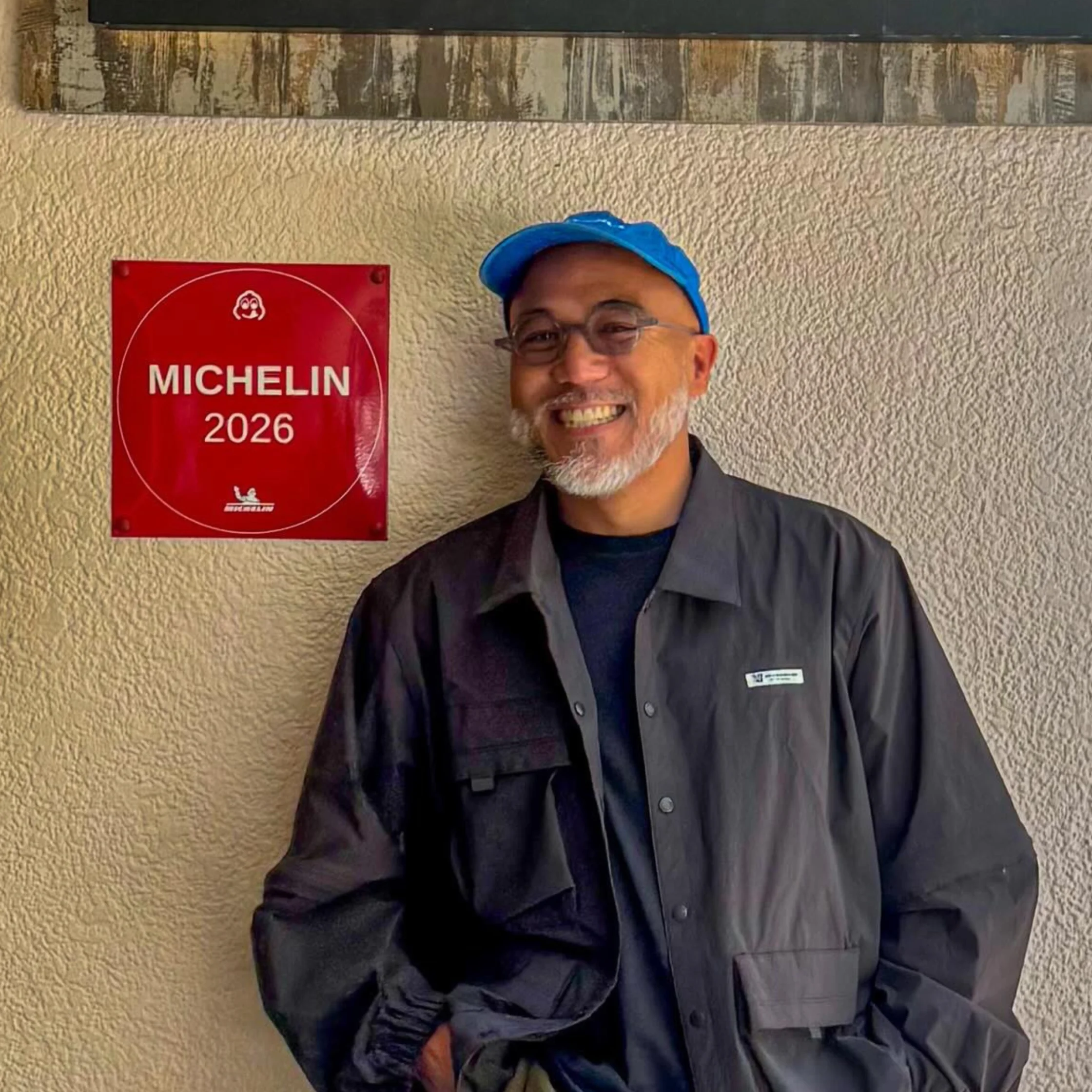 Smiling restaurant worker in a blue cap and dark uniform stands beside a red MICHELIN 2026 plaque on a textured wall, conveying pride and warmth.