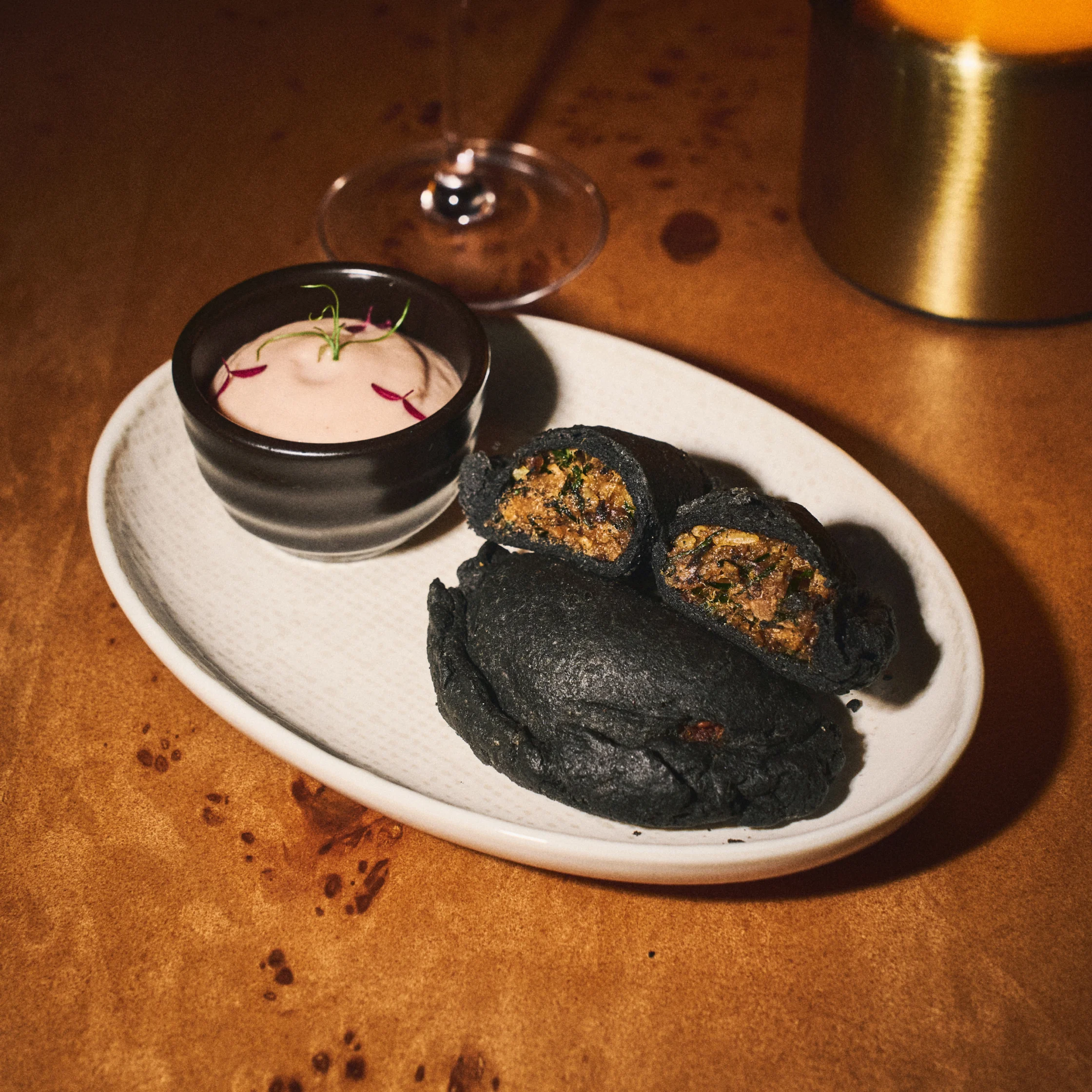 A white plate holds charcoal-black buns, two cut open to reveal a savory filling, beside a bowl of pale dipping sauce; a wine glass adds a warm, intimate restaurant mood.