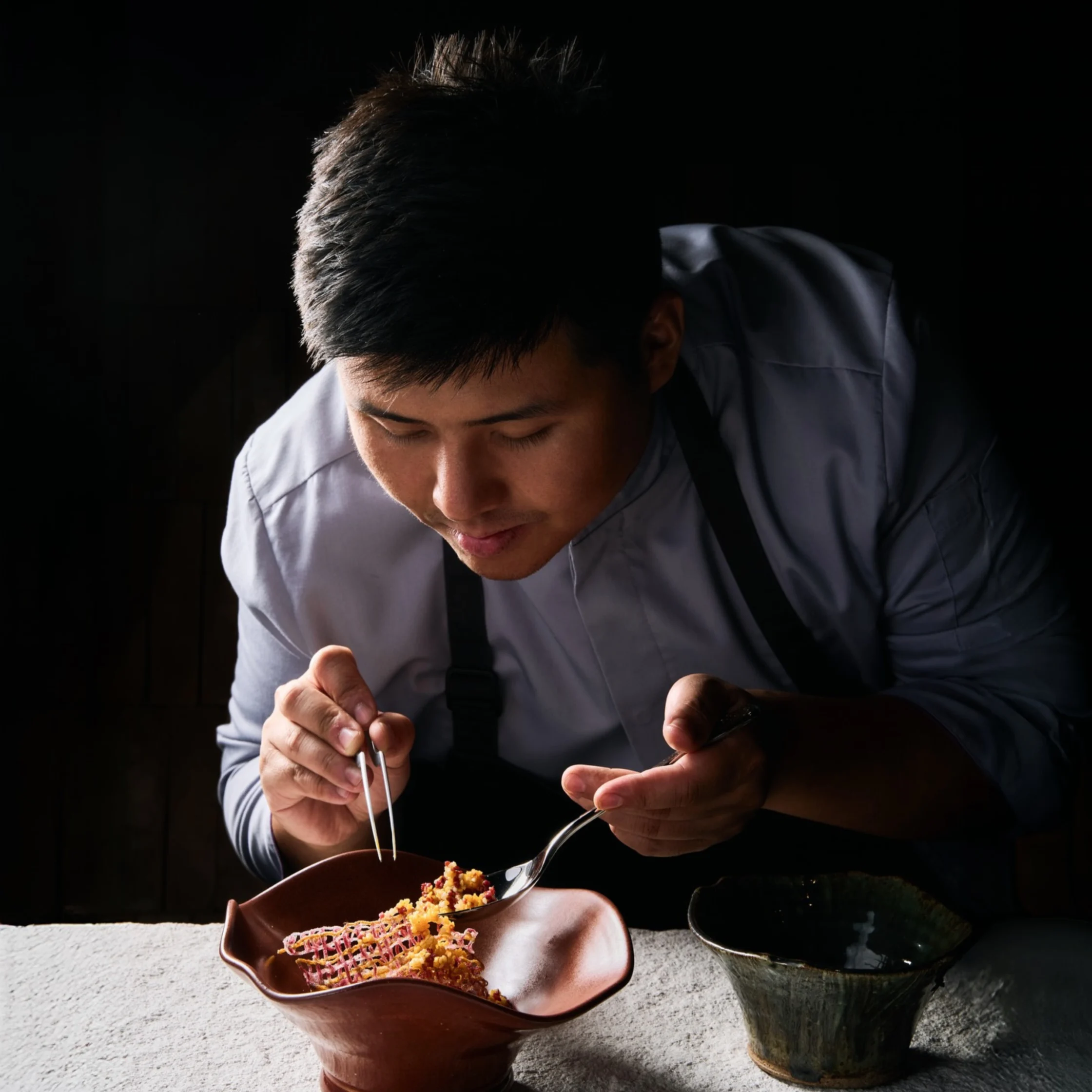 A young chef in a white jacket leans over a bowl, carefully tasting a colorful dish with a fork and spoon in dramatic, low-key lighting.