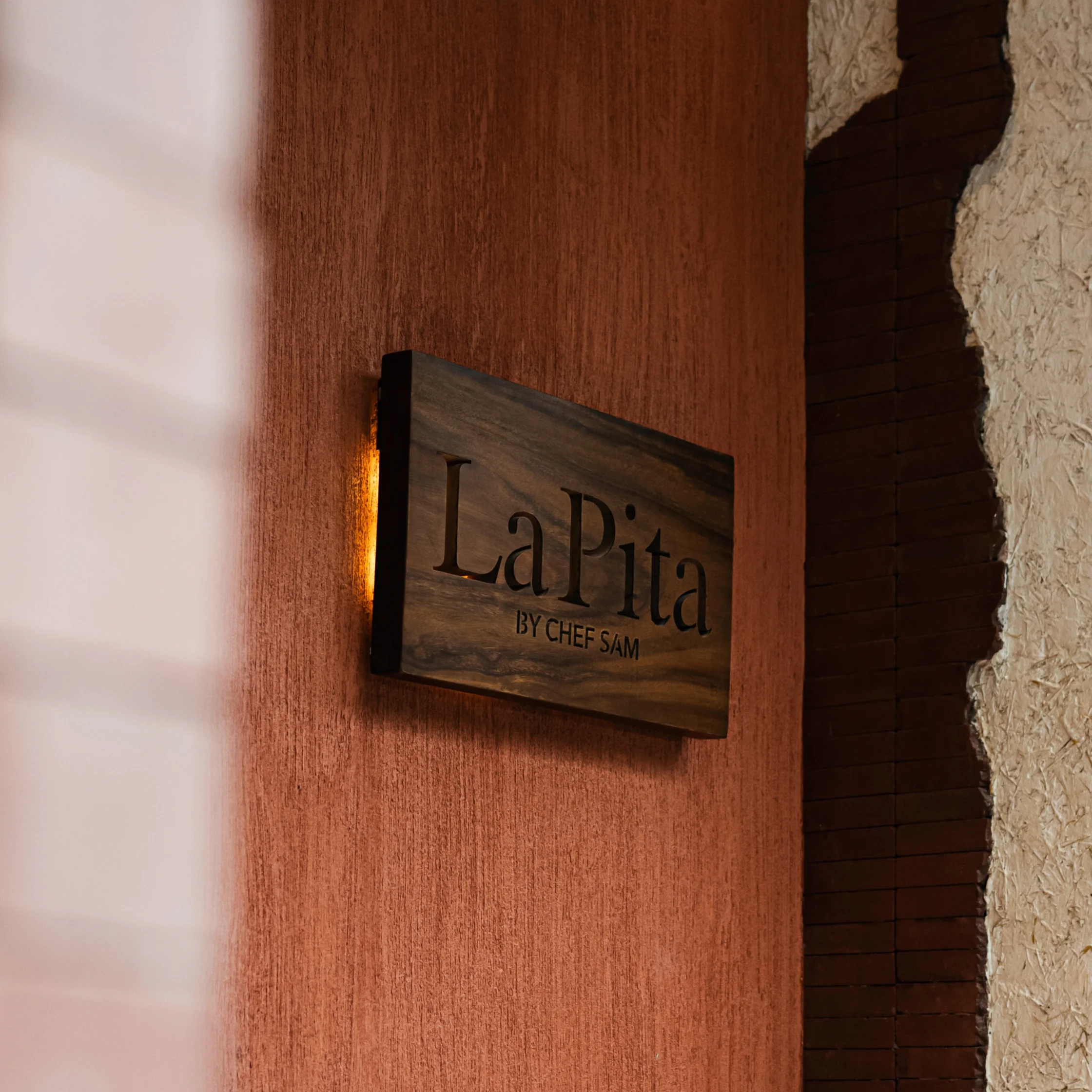 A wooden “LaPita” restaurant sign hangs on a terracotta wall beside a rough stone doorway, creating a warm, rustic and inviting atmosphere.
