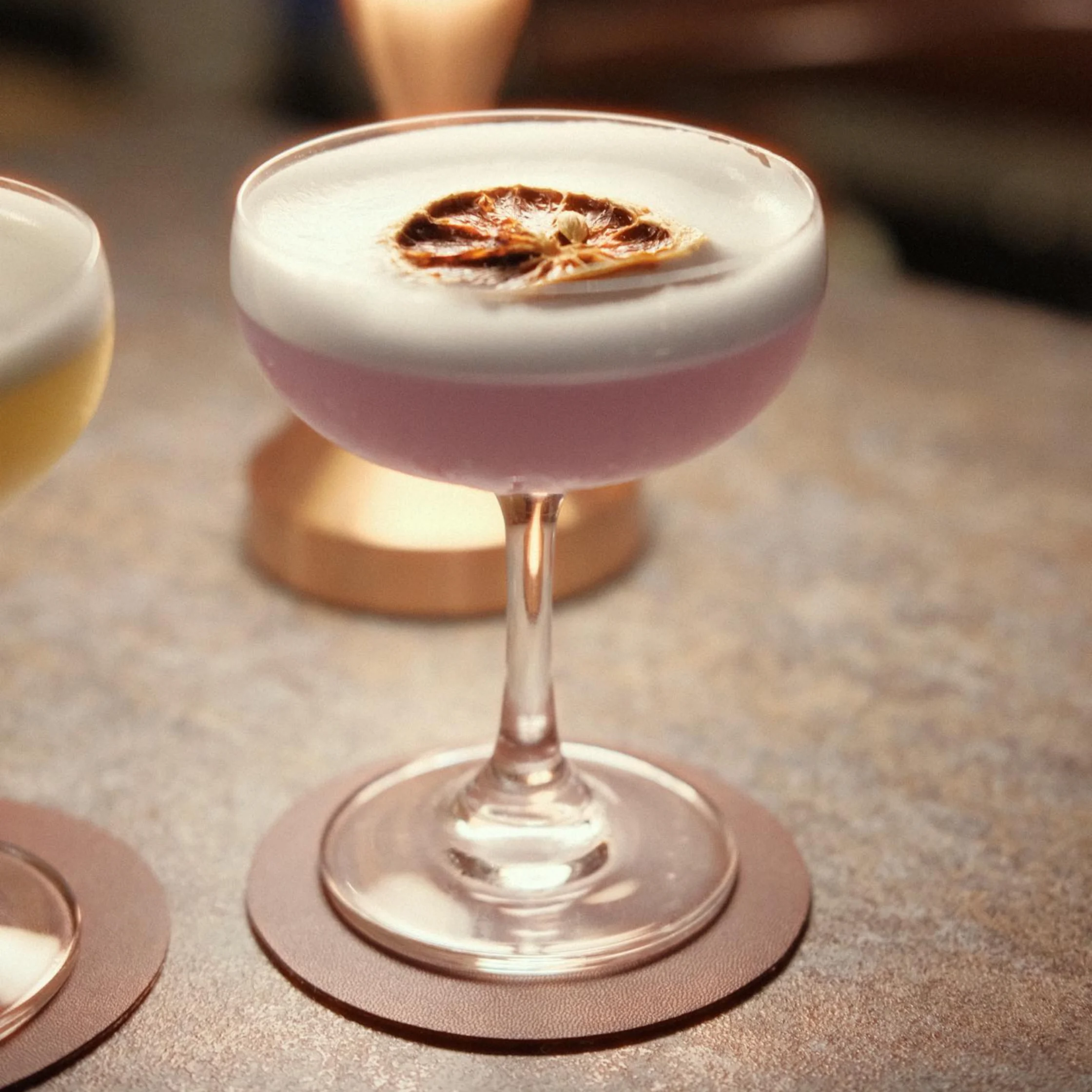 A pink cocktail with a thick white foam cap and dried citrus garnish sits in a coupe glass on a coaster, warmly lit in a cozy bar.