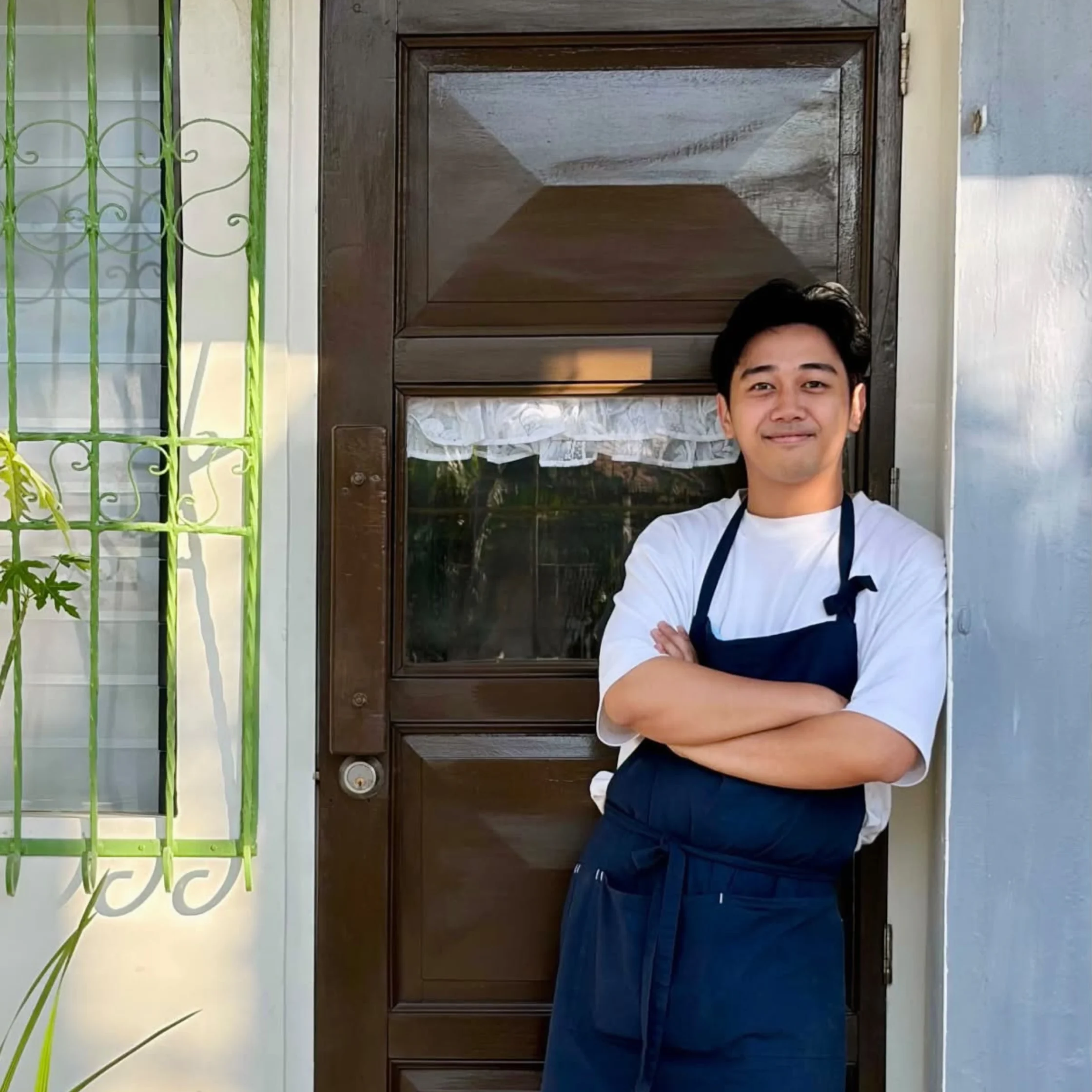 A smiling young man in a navy apron stands with arms crossed outside a dark wooden door, beside a green barred window, creating a calm, welcoming mood.