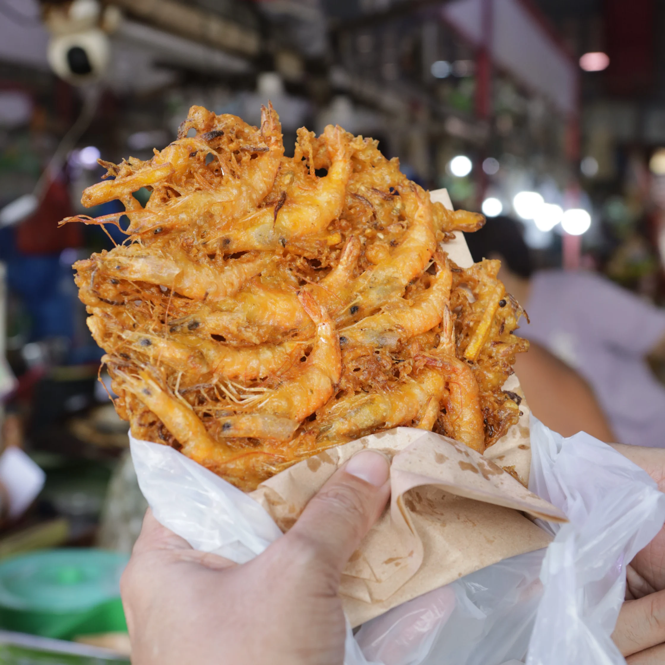 A hand holds a paper-lined bag overflowing with golden, crispy fried shrimp at a busy night market, highlighting the tempting street-food snack.