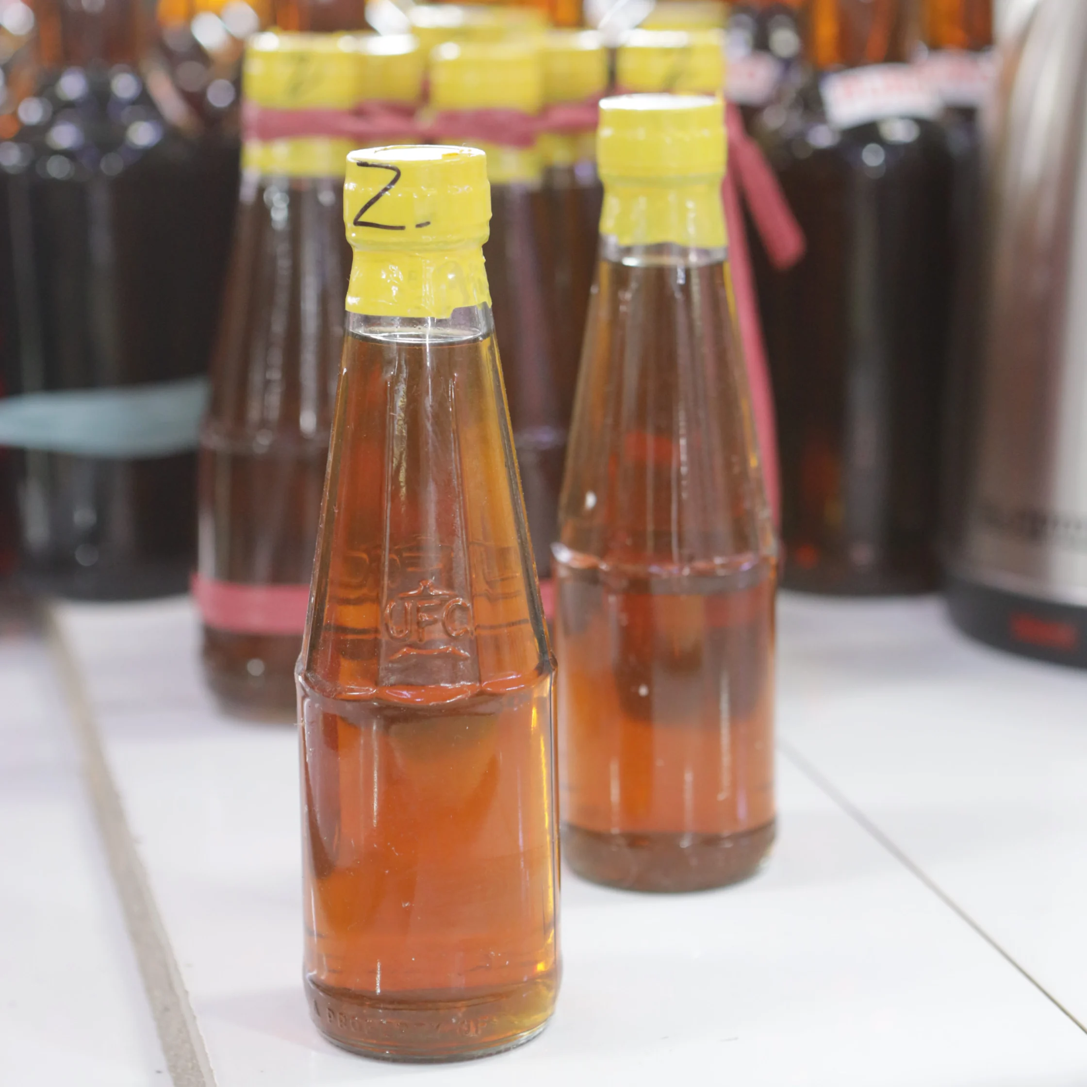 A clear glass bottle of amber-colored sauce with a yellow cap stands on a white counter among rows of condiments, creating a busy shop display.