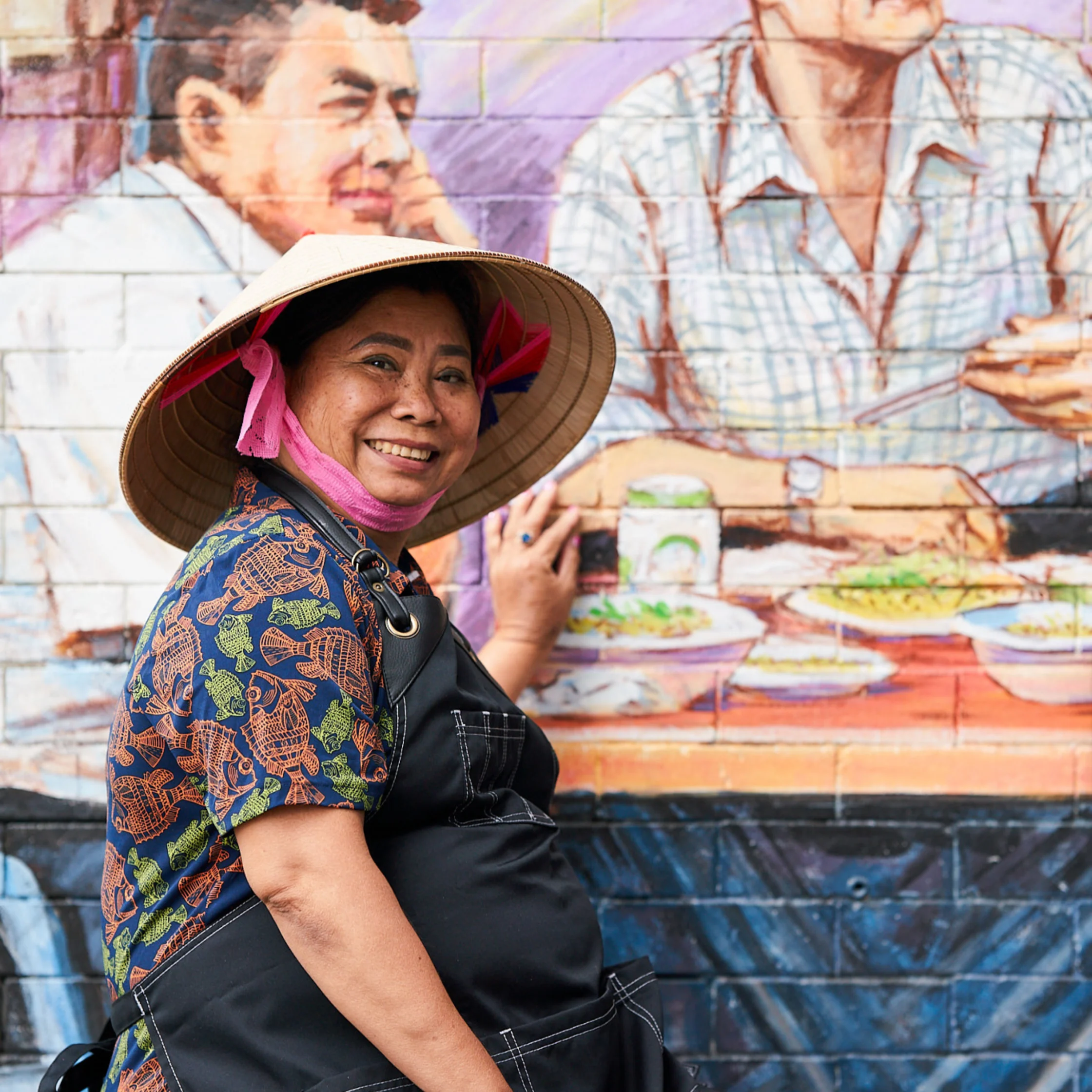 A smiling woman in a conical hat and patterned shirt poses beside a colorful Vietnamese street-food mural, creating a warm, lively scene.