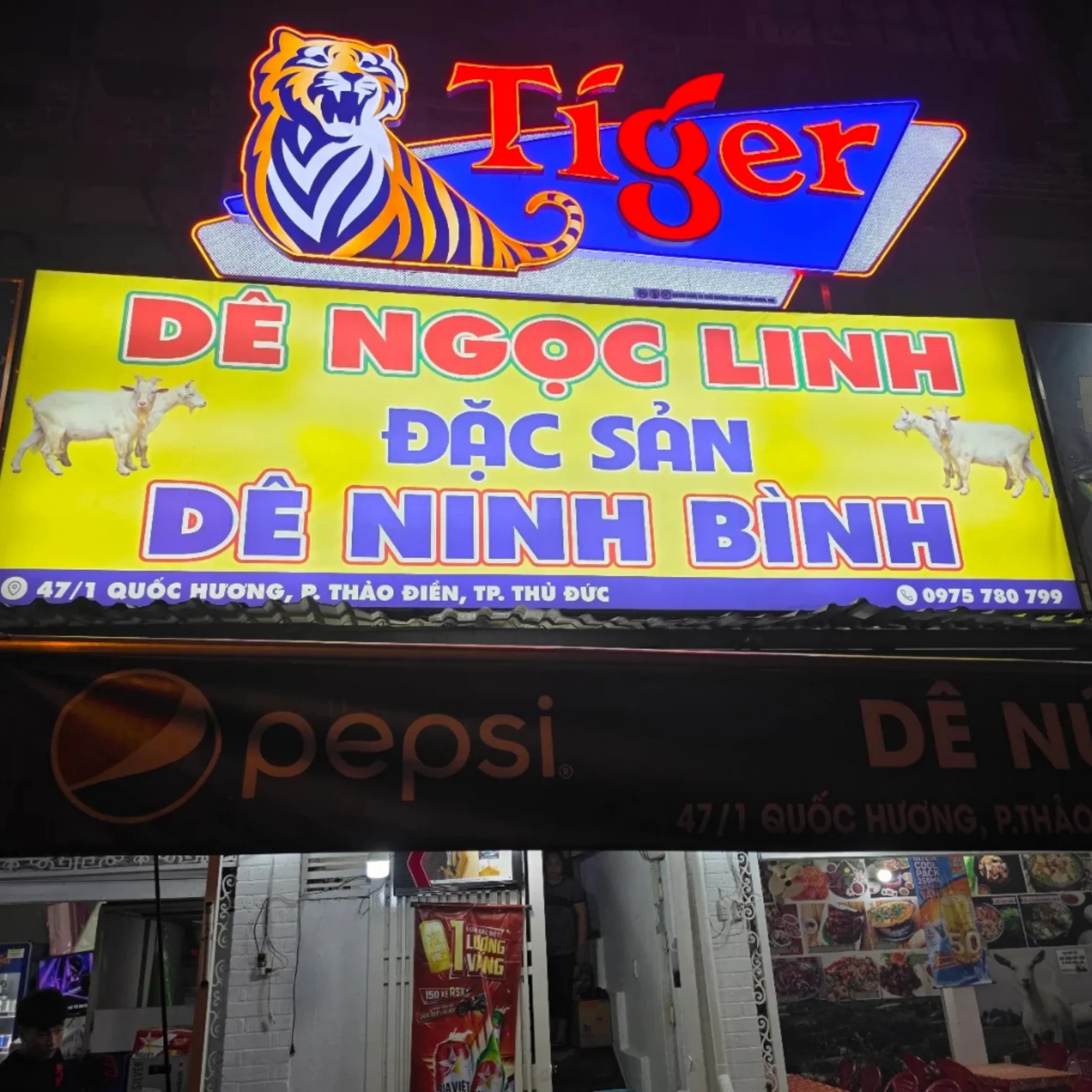 A brightly lit Vietnamese restaurant sign reads “Tiger Dê Ngọc Linh,” advertising Ninh Bình goat specialties with tiger and goat illustrations. Below, a Pepsi-branded awning gives the nighttime street scene a colorful, lively feel.