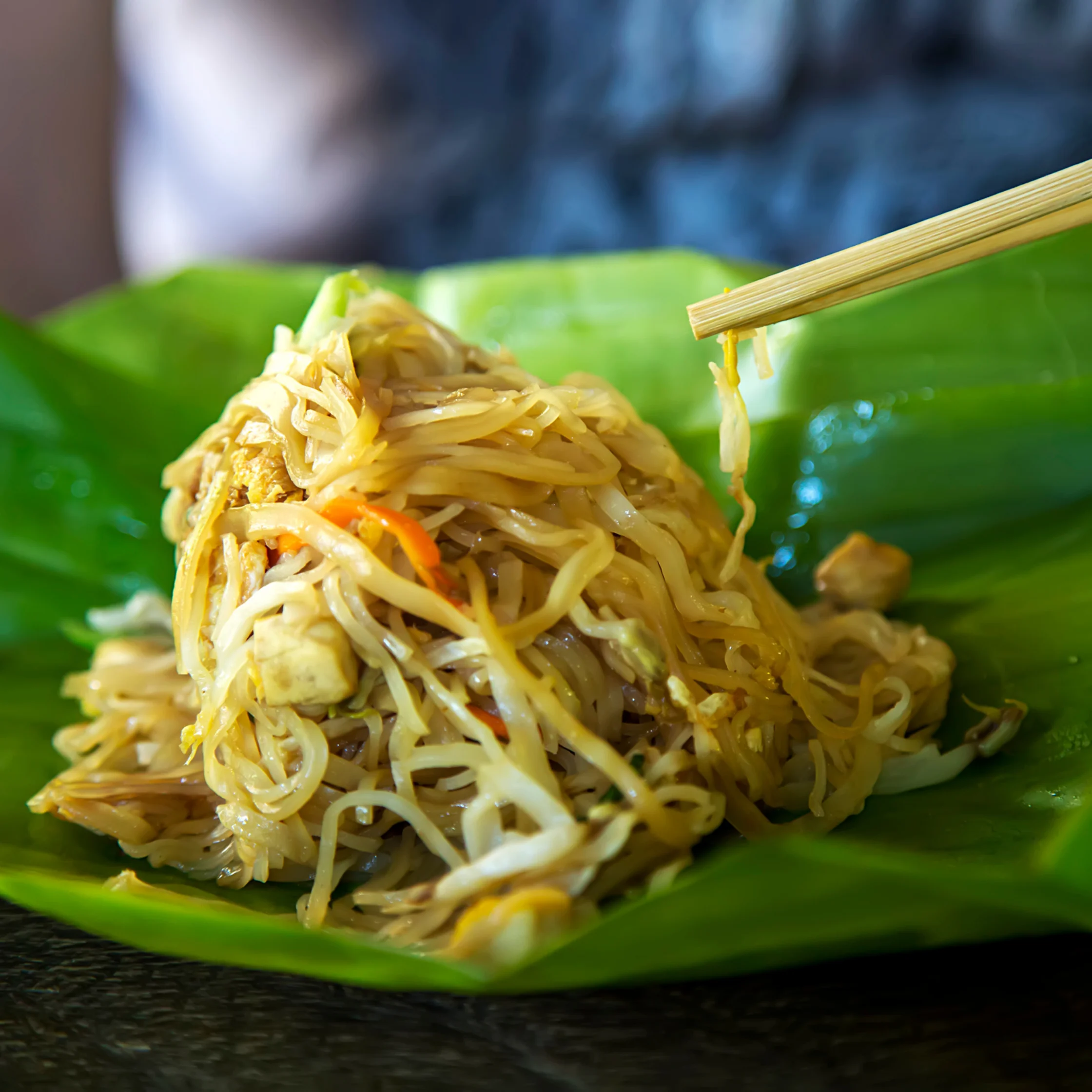 A glossy mound of stir-fried noodles with vegetables sits on a bright green banana leaf, while chopsticks lift a strand; appetizing, vibrant close-up.