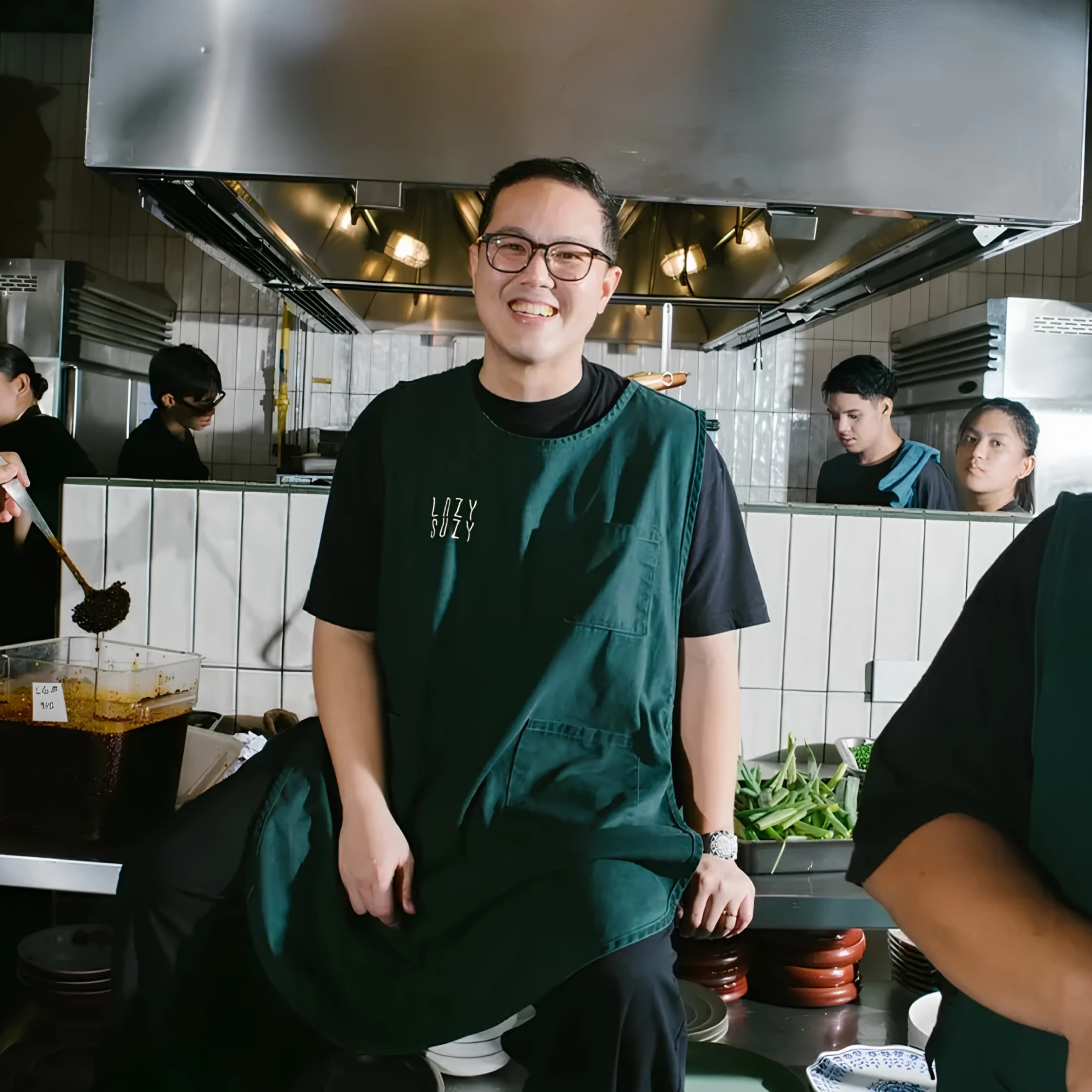 A smiling cook in glasses and a dark green apron stands in a busy restaurant kitchen, with staff, cooking equipment, and fresh greens behind him; warm, welcoming atmosphere.