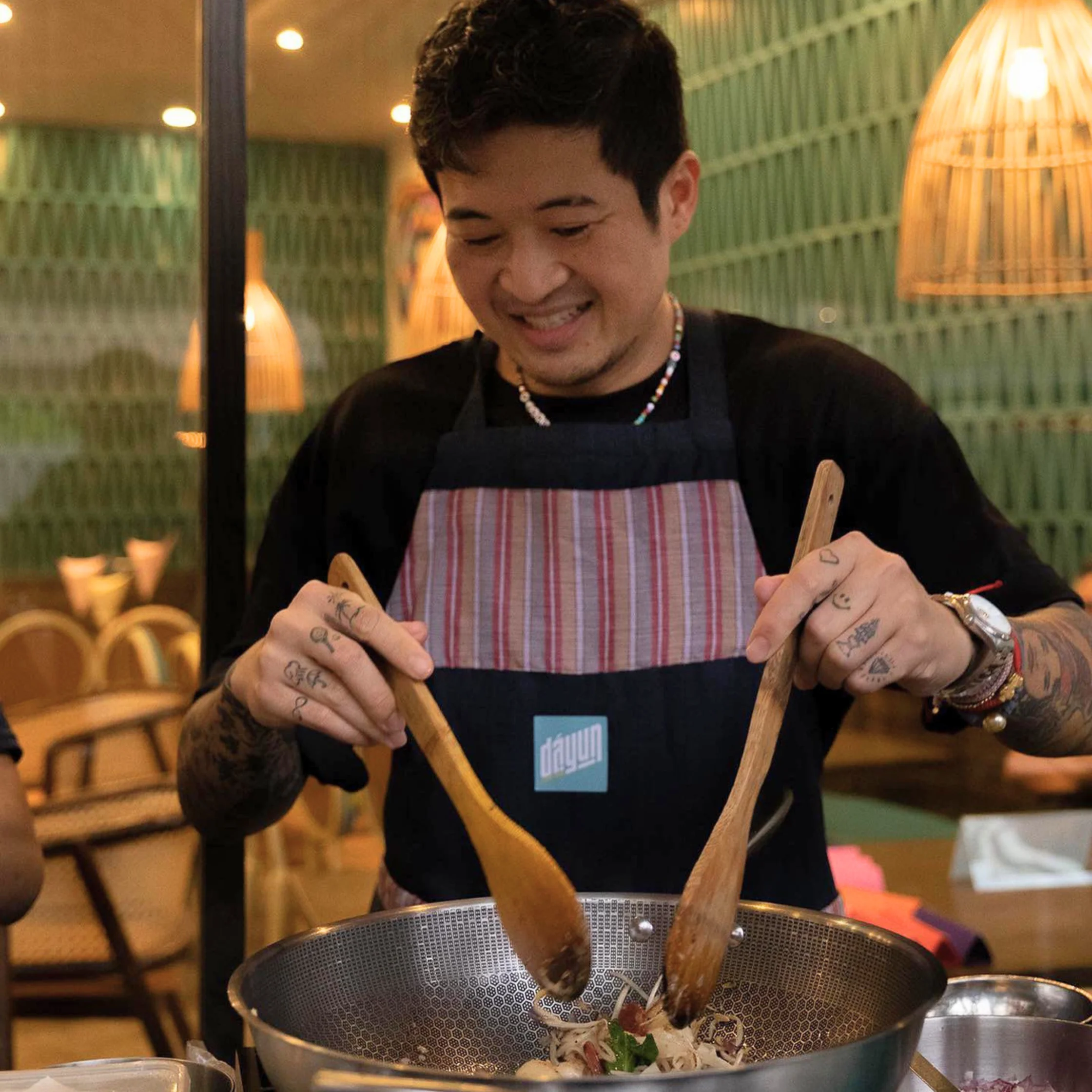 A smiling tattooed cook in a striped apron stirs ingredients in a large metal wok at a warmly lit restaurant, creating a friendly, focused mood.