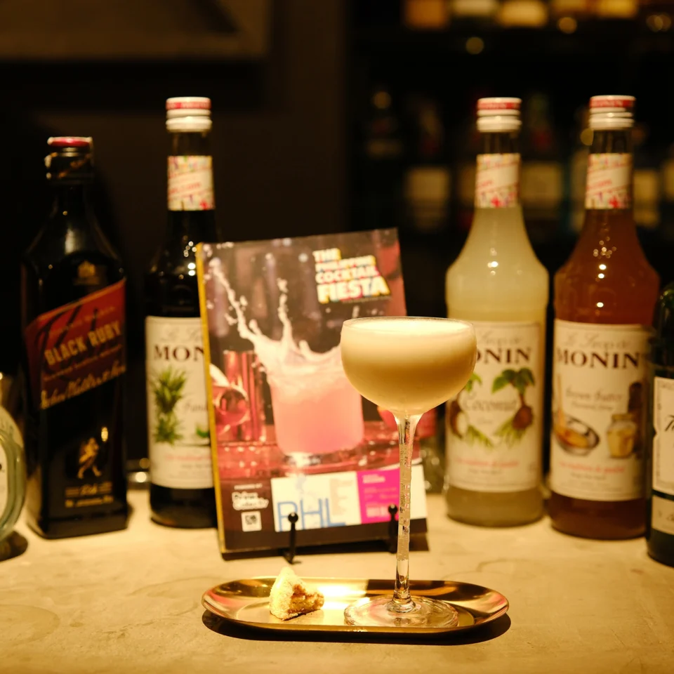 A frothy pale-yellow cocktail in a stemmed glass sits on a tray with a garnish, surrounded by liquor and Monin syrup bottles and a cocktail menu, in warm, inviting bar light.