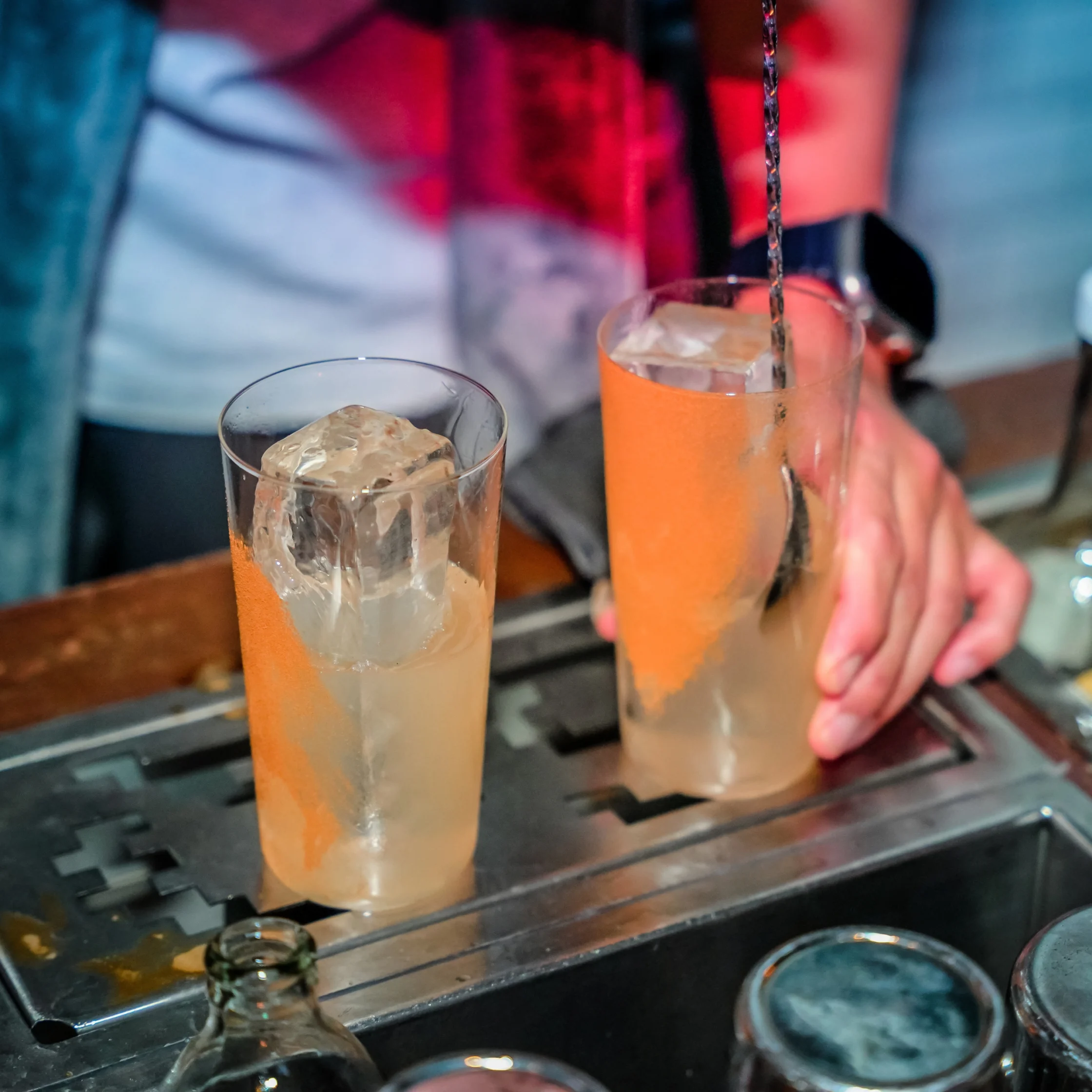 A bartender stirs an orange iced cocktail in a tall glass beside a second drink on a metal bar, capturing the lively craft of drink preparation.