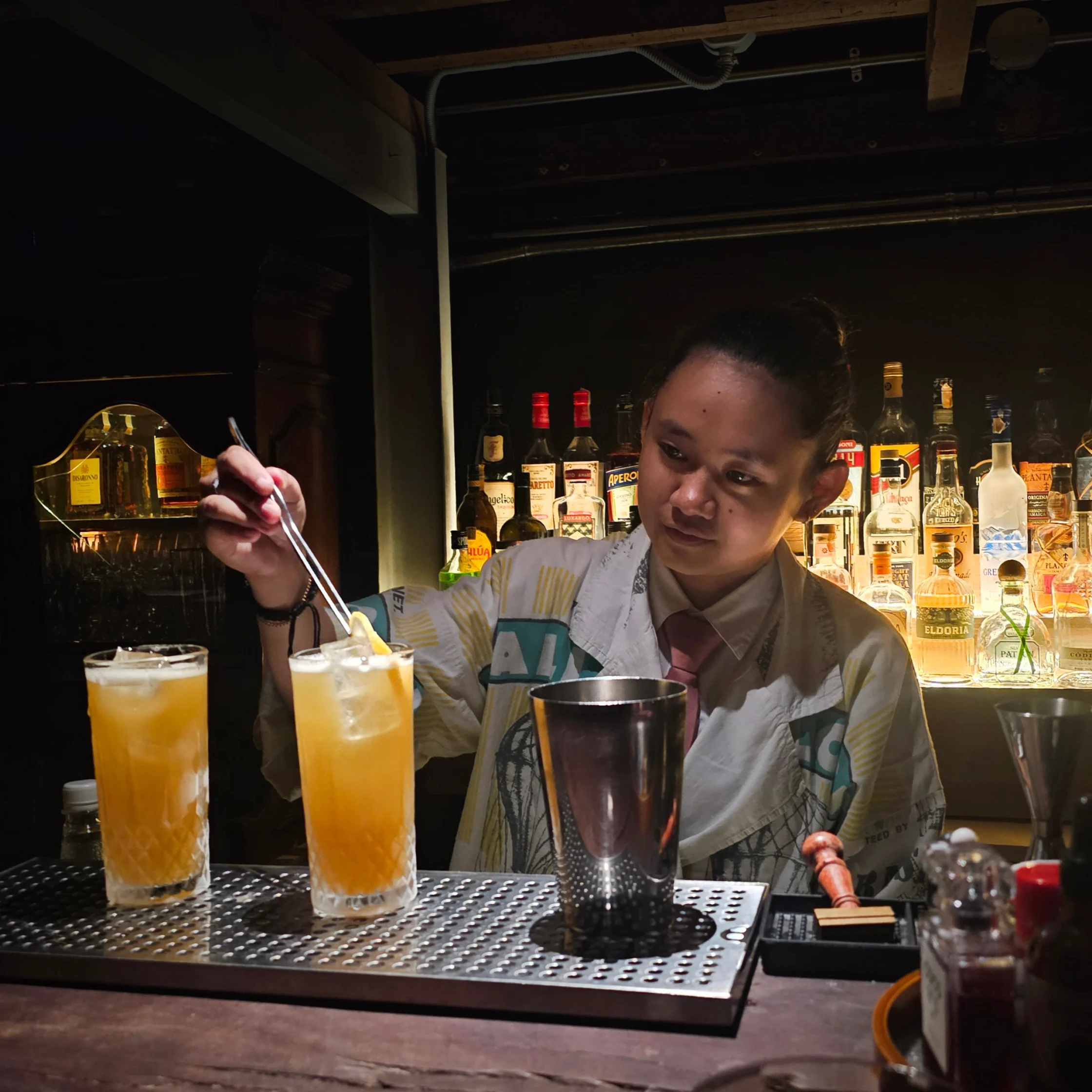 A focused bartender stirs two tall orange cocktails behind a metal bar, with liquor bottles glowing on shelves in the dim, intimate setting.