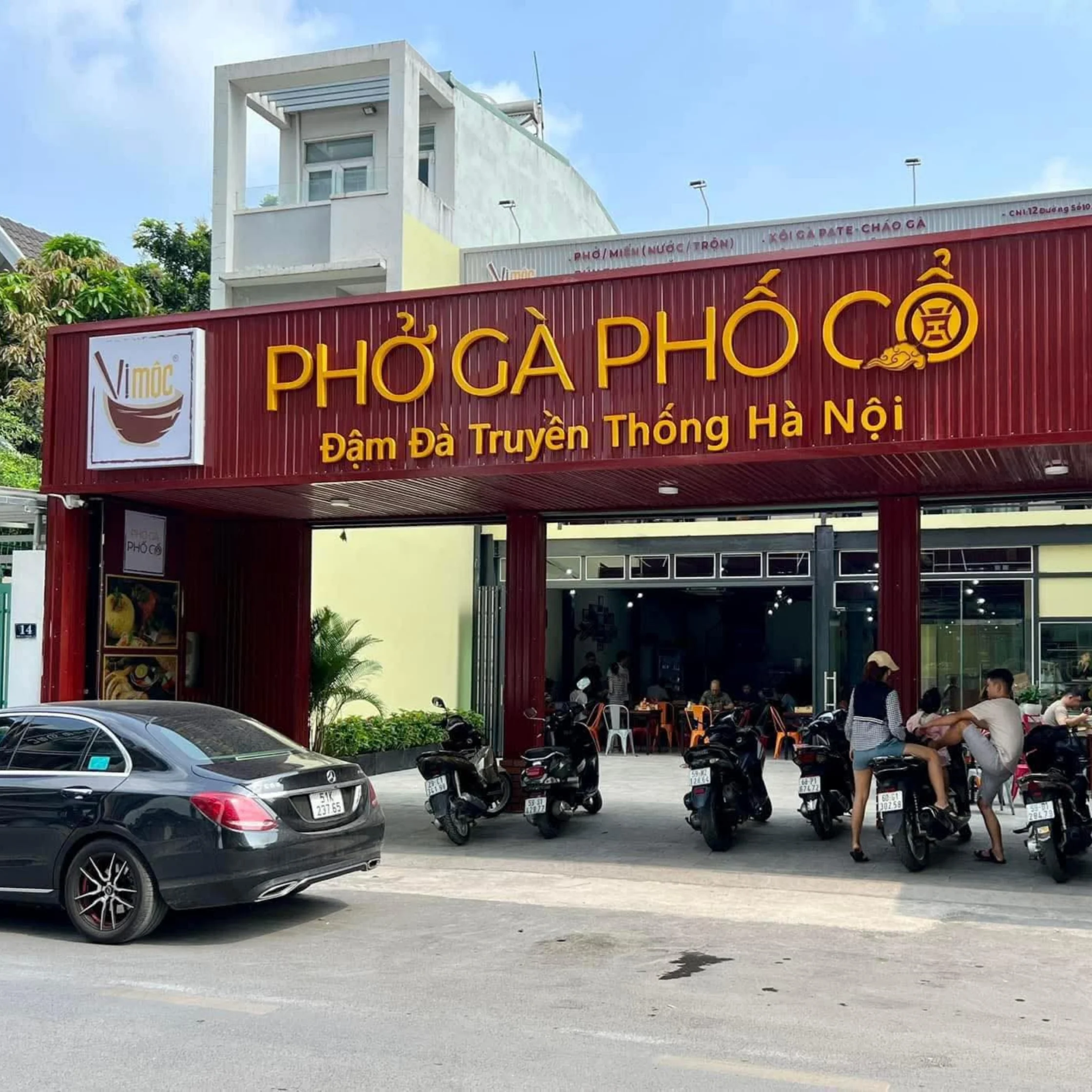 A busy Hanoi pho restaurant with a large red sign reading “Phở Gà Phố Cổ,” parked motorbikes, a car, and diners outside in bright daylight.