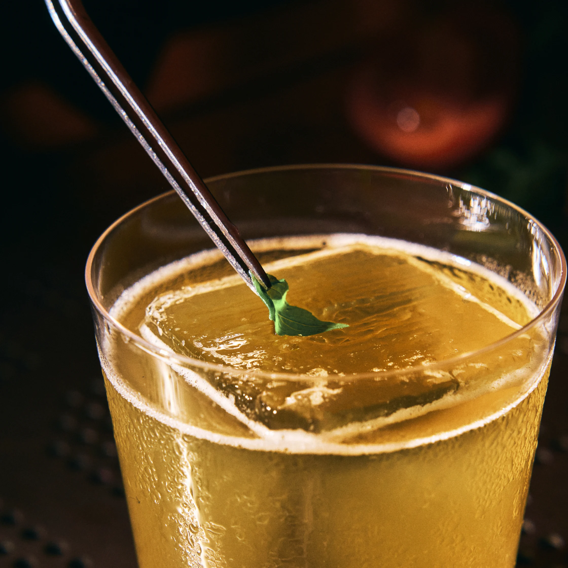 Close-up of a chilled golden cocktail in a clear glass, with ice and a metal pick holding a small green garnish; warm, moody bar lighting creates an inviting tone.