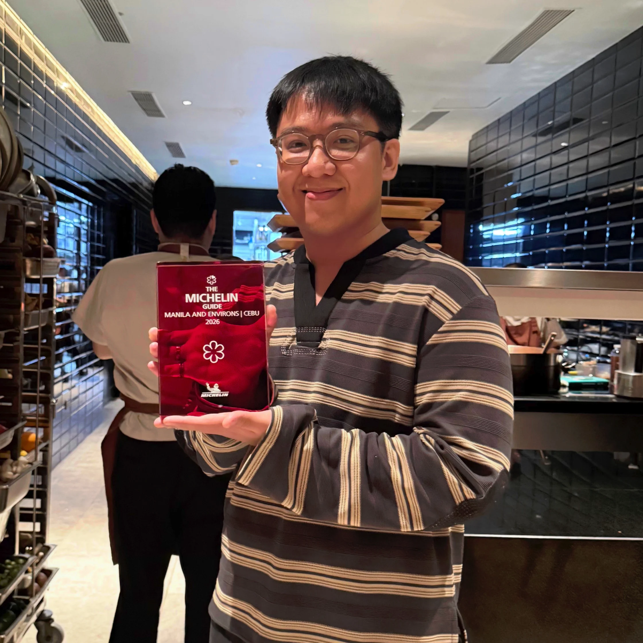A smiling man in glasses holds a red Michelin Guide award plaque inside a modern restaurant kitchen, conveying pride and celebration.