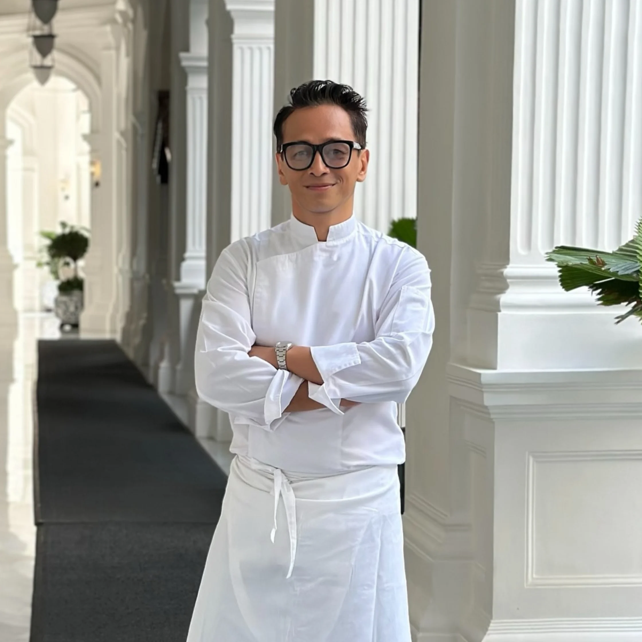 A confident chef in a white jacket and apron stands with arms crossed in a bright, elegant colonnade, smiling calmly at the camera.