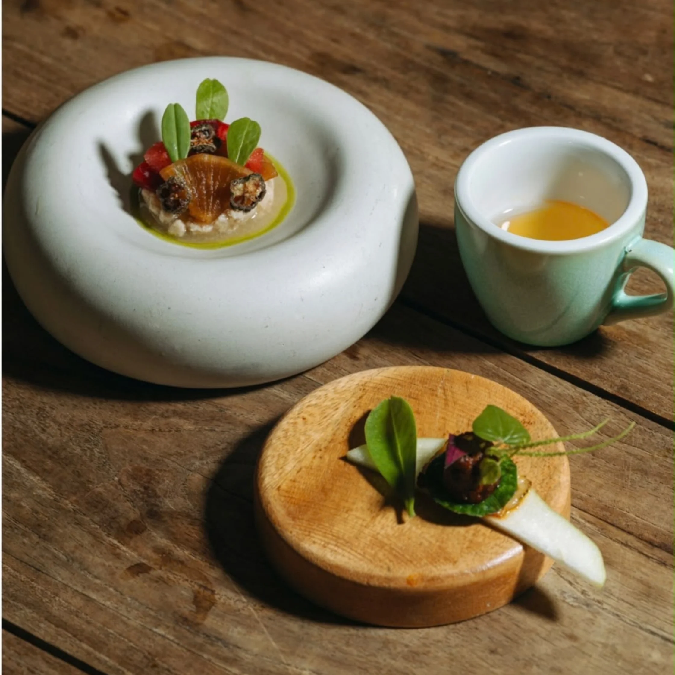 Three artfully plated gourmet bites with leafy greens and colorful garnishes sit on ceramic dishes beside a cup of golden tea, on a rustic wooden table.