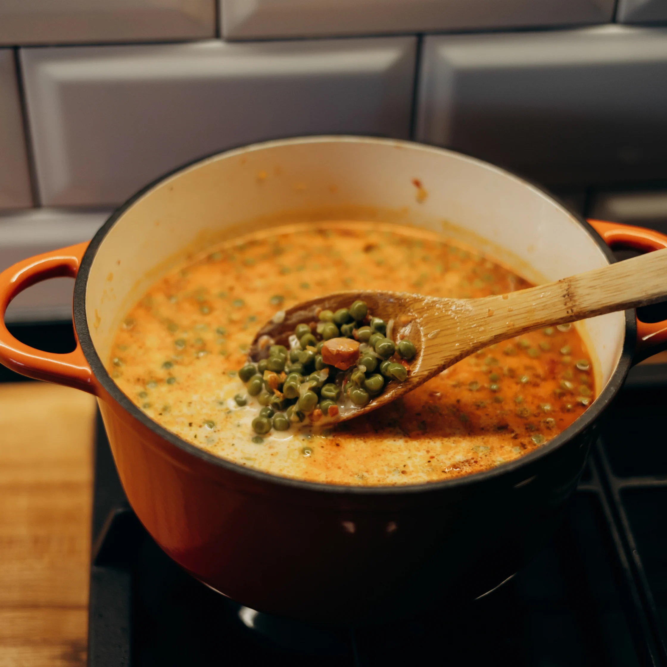 A wooden spoon lifts green peas and diced vegetables from a creamy orange soup simmering in an orange Dutch oven on the stove, creating a warm, cozy kitchen mood.