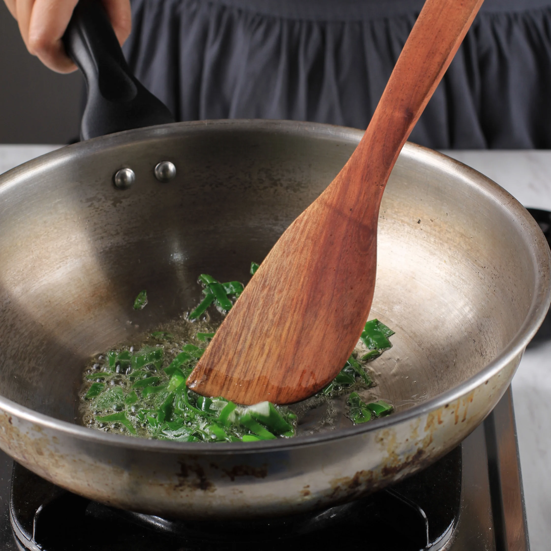 A cook stirs chopped green herbs in a well-used metal pan on a stovetop with a wooden spatula; a warm, hands-on kitchen scene.