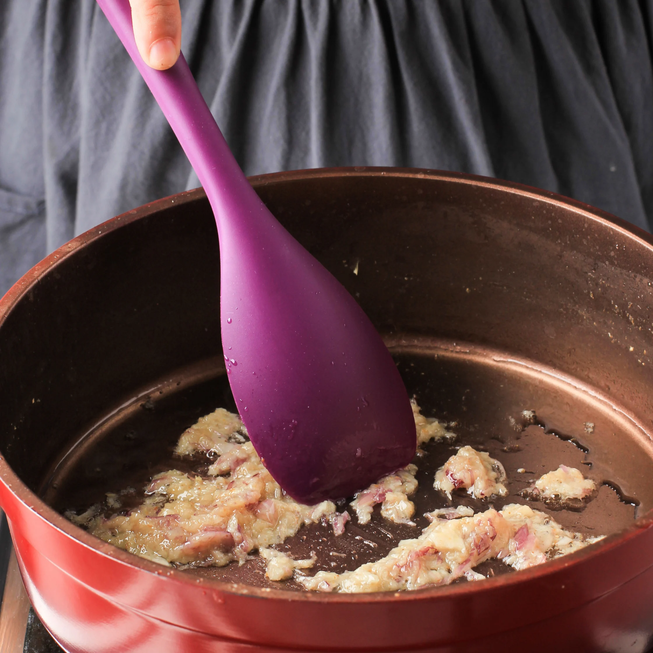 A hand stirs chopped onions and bacon in a red frying pan with a purple spatula, suggesting a warm, hands-on cooking scene.