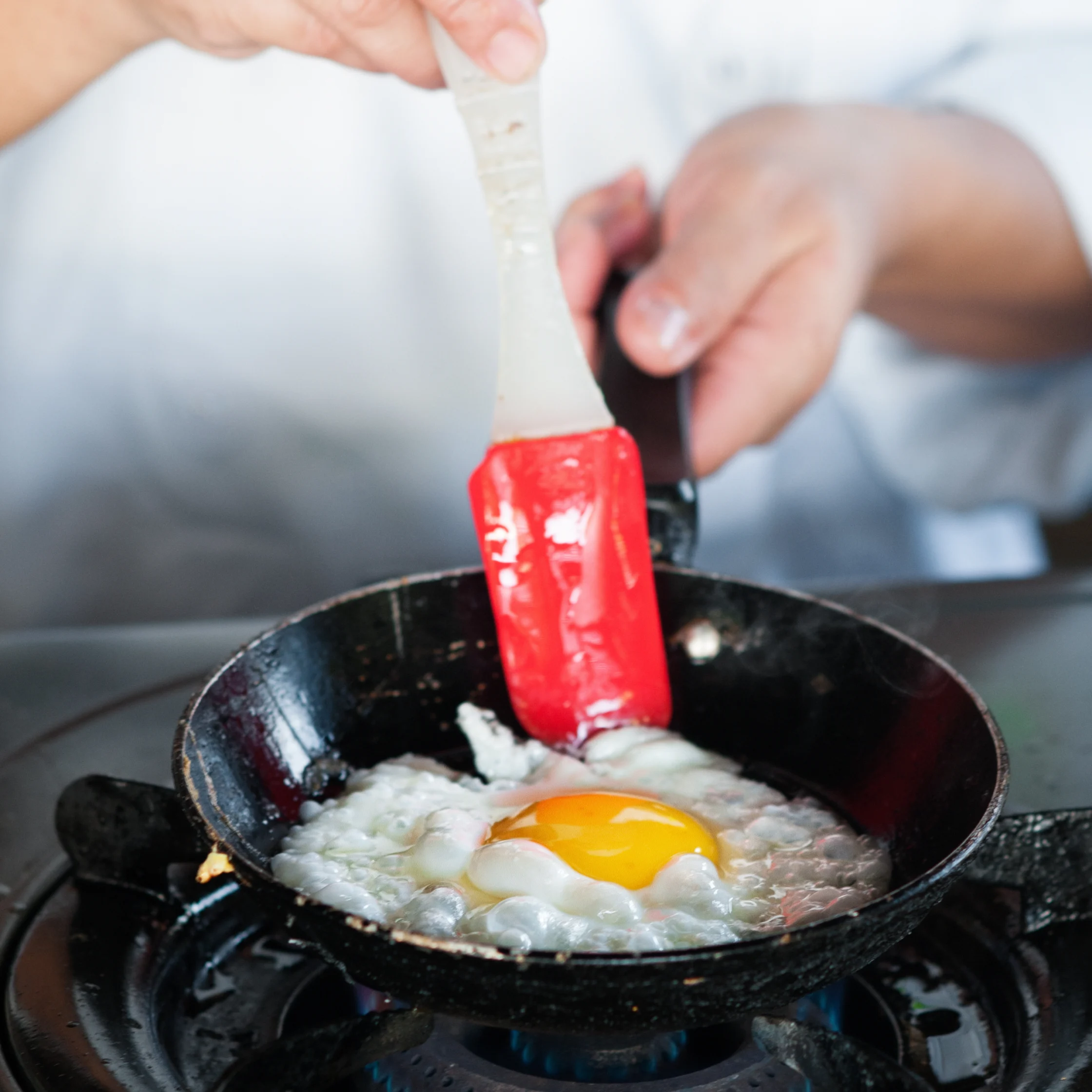 A cook uses a red spatula to gently lift a sunny-side-up egg in a black frying pan over a gas flame, creating a warm, homey kitchen scene.