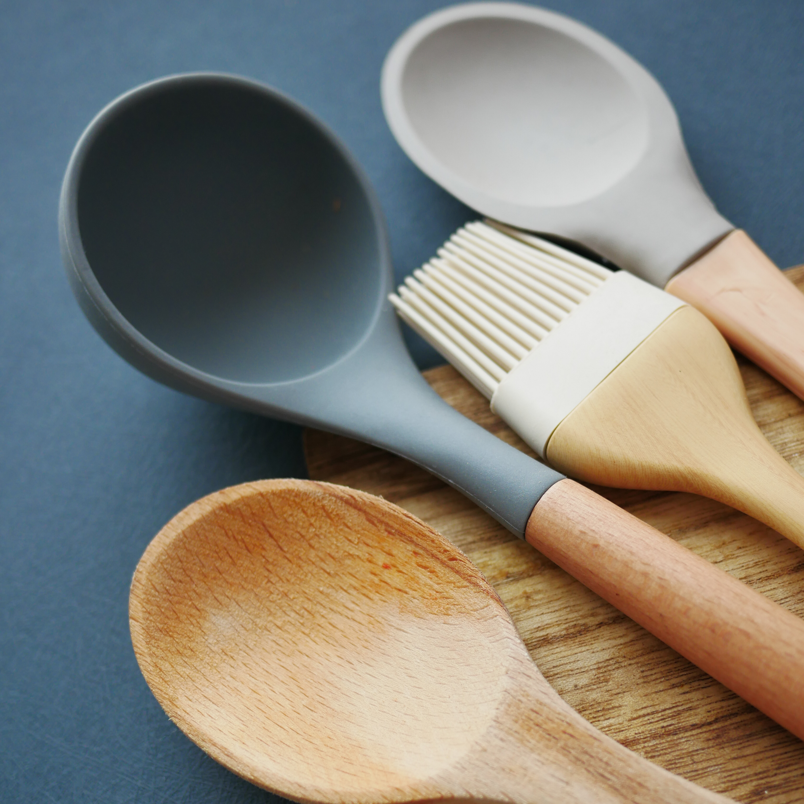A neat arrangement of wooden cooking spoons, a gray ladle, pastry brush, and white spatula on a blue surface, evoking a calm, practical kitchen.