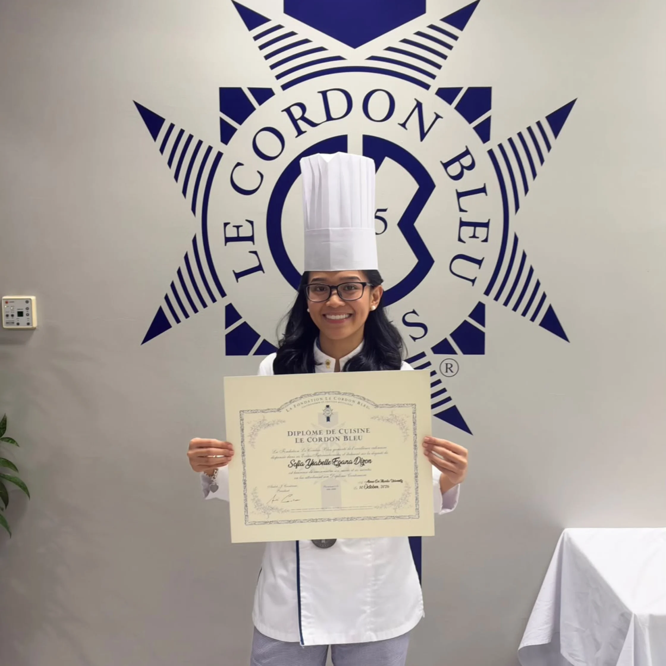 A smiling chef in a tall white toque and uniform holds a culinary diploma before a Le Cordon Bleu logo, celebrating her achievement.