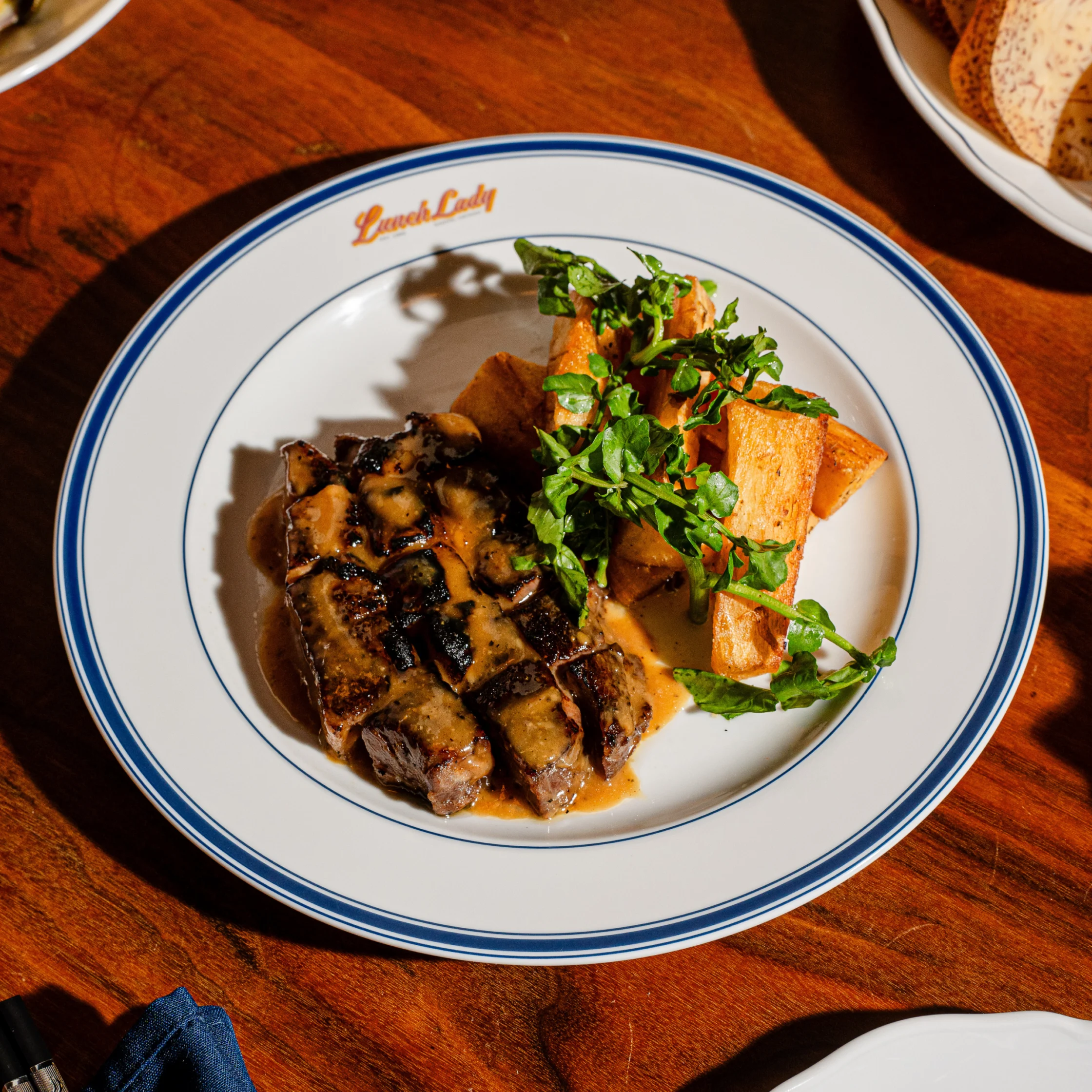 Grilled steak and golden roasted potatoes topped with fresh parsley on a blue-rimmed plate, presented in warm sunlight on a wooden table.