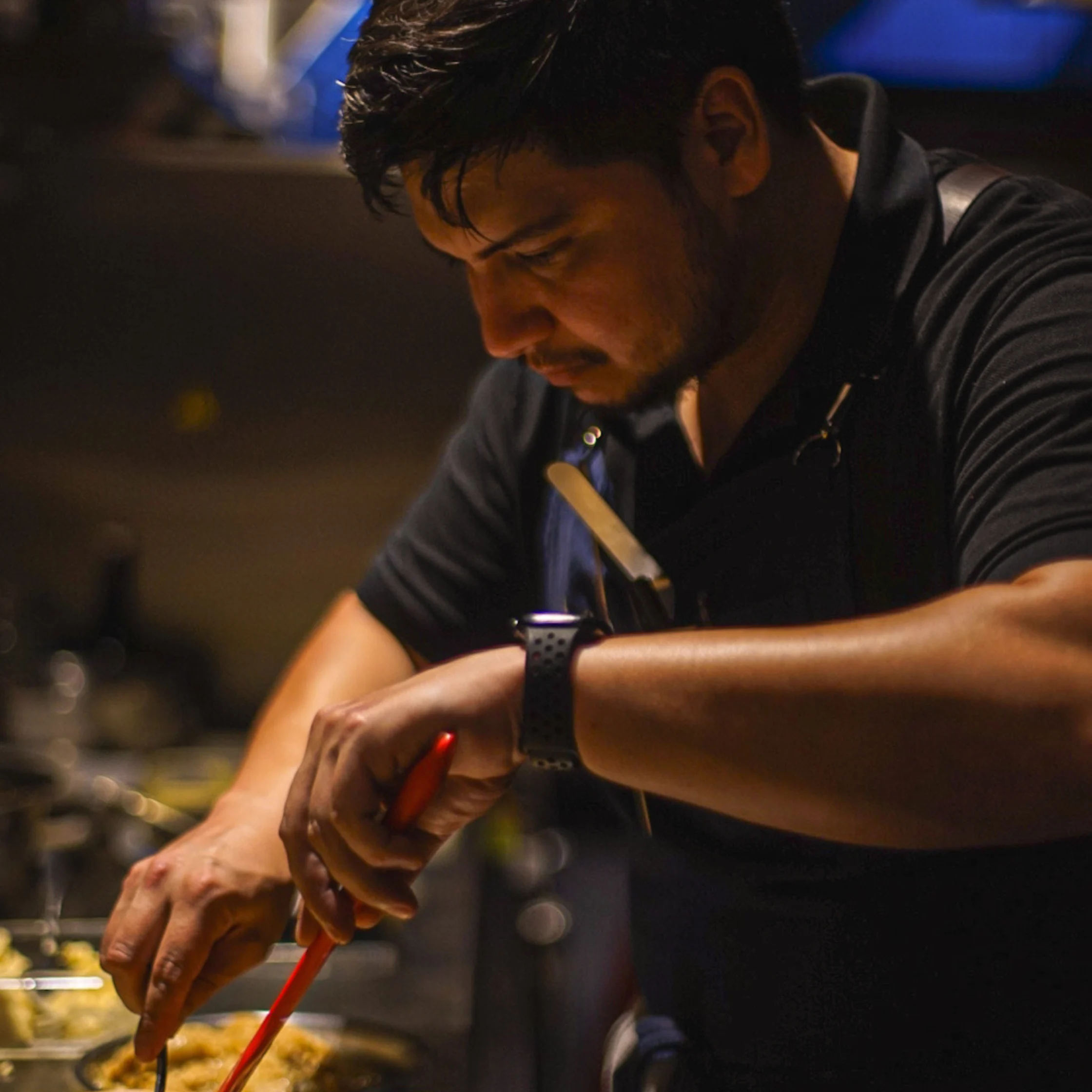 A focused chef in a dark shirt leans over a sizzling pan, stirring food with red chopsticks in a dimly lit kitchen; warm, intense atmosphere.