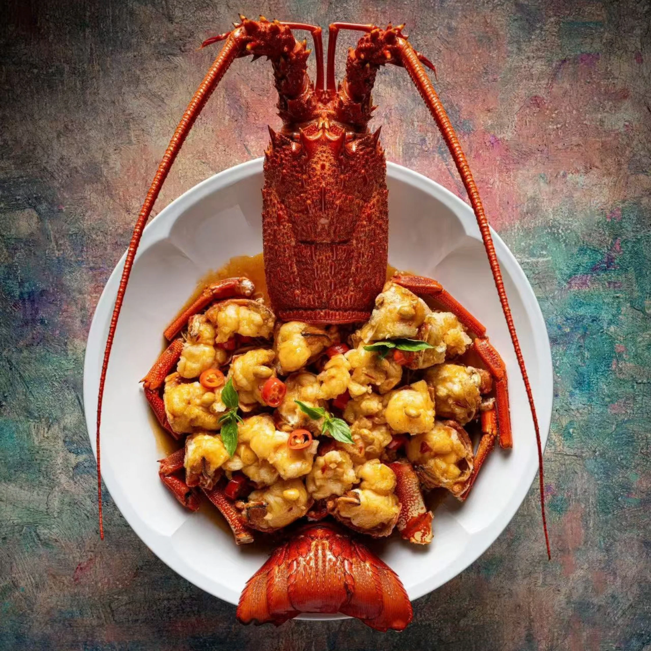 A cooked lobster, antennae extended, sits on a white plate filled with golden seafood pieces and herbs, creating a rich, celebratory dining scene.