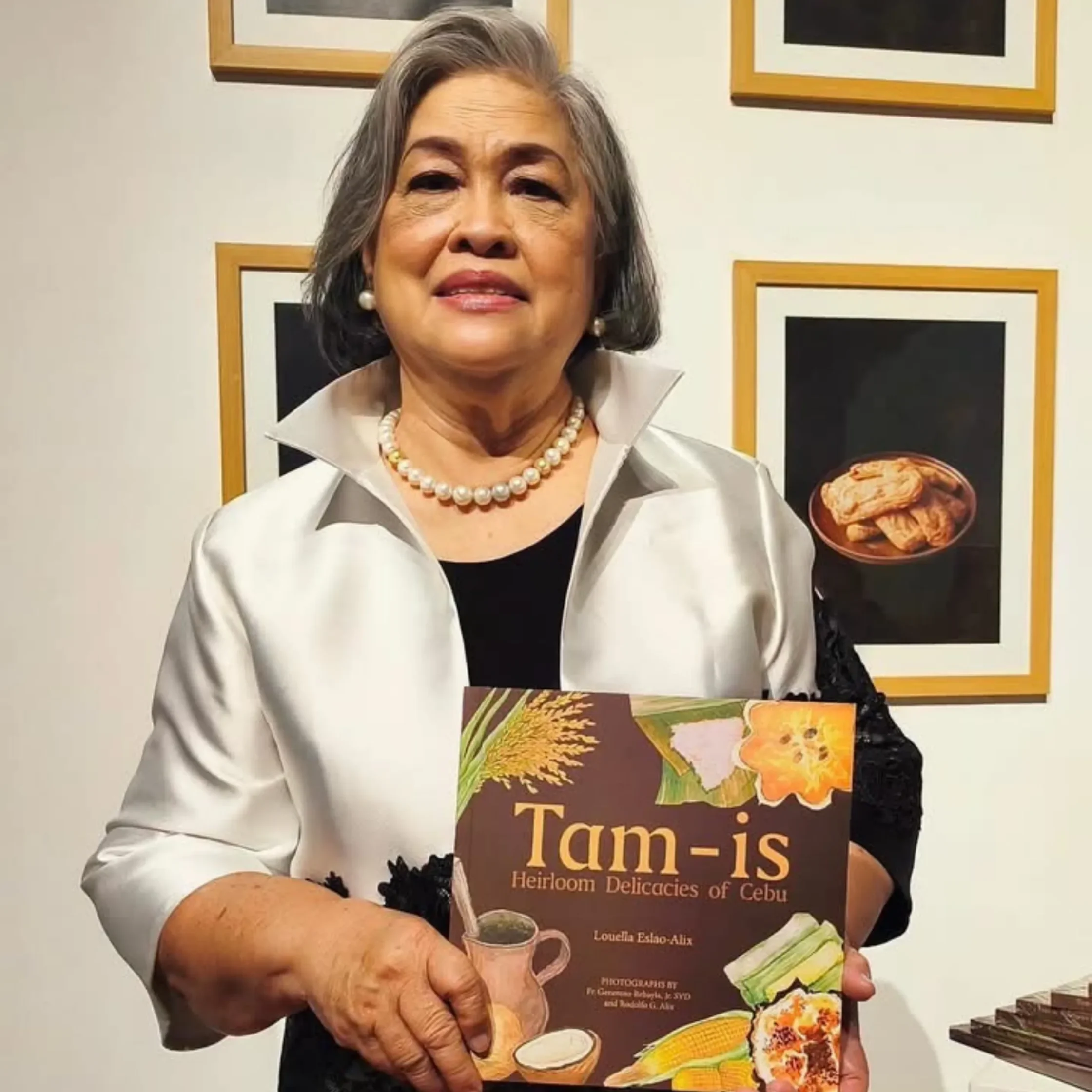 An older woman smiles in a gallery, holding “Tam-is,” a cookbook about Cebu delicacies; framed food art surrounds her, creating a warm, welcoming tone.