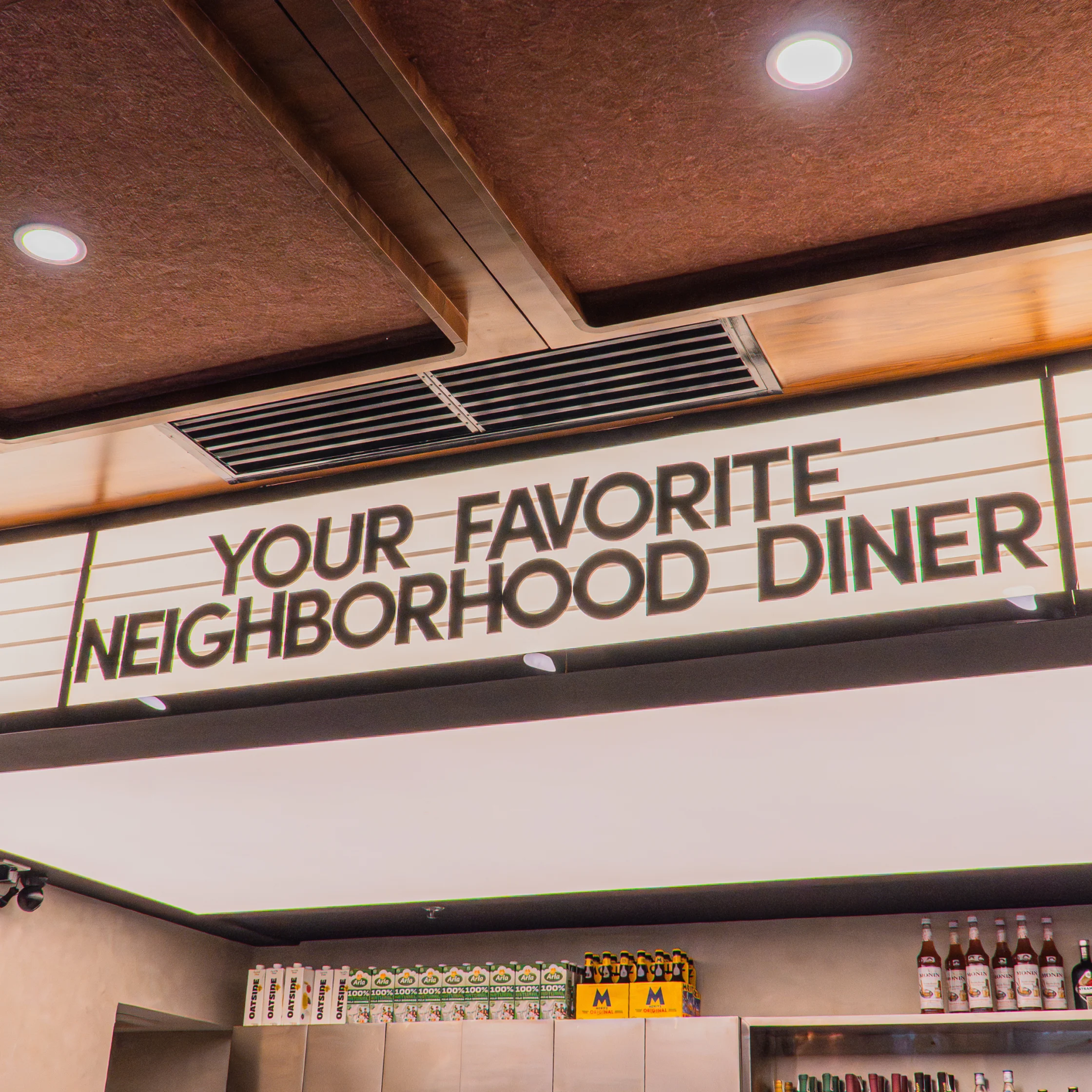 An illuminated diner sign reading “Your Favorite Neighborhood Diner” hangs beneath a warm, textured ceiling, with shelves of drinks below; cozy, nostalgic mood.