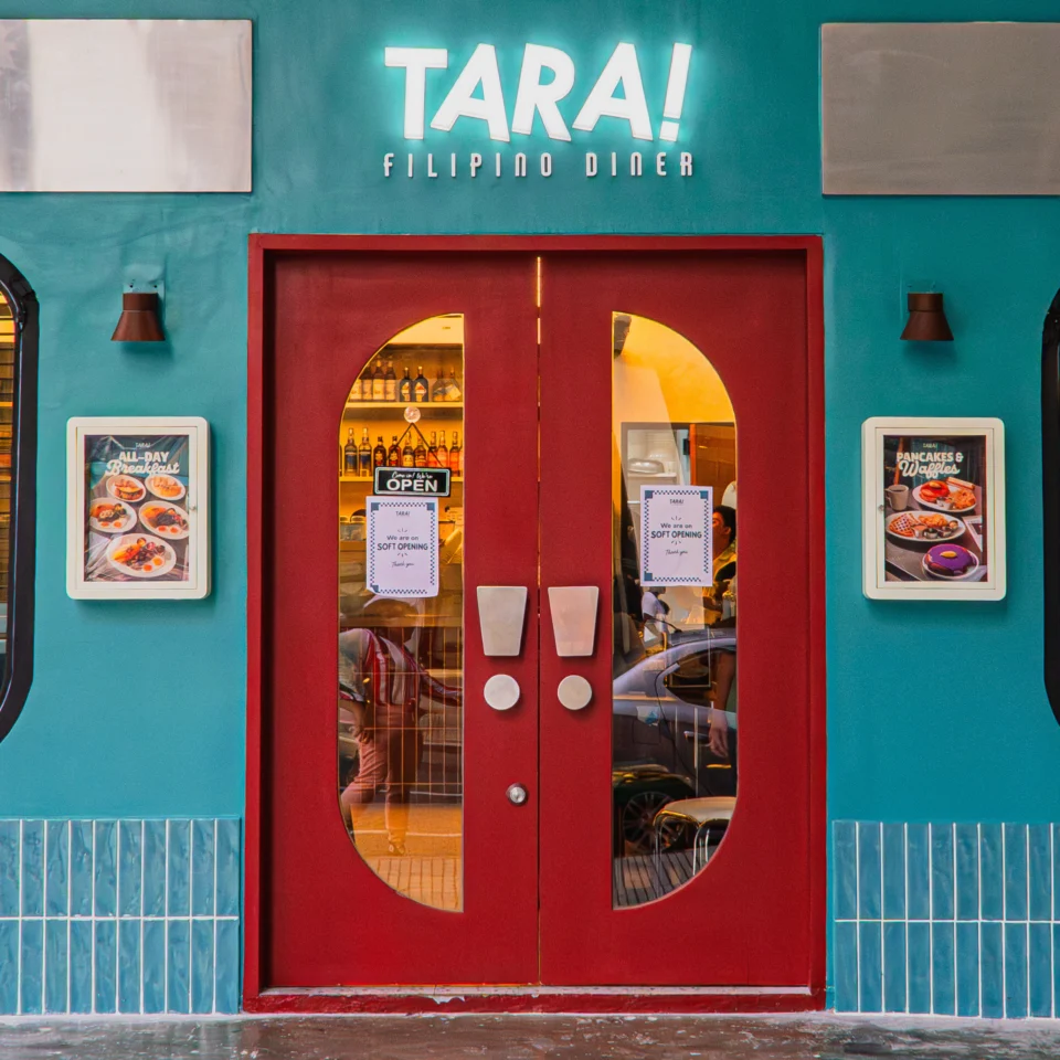 Teal Tara! Filipino diner storefront with bright red double doors, glowing sign, food photos, and window lettering advertising breakfast and coffee; inviting feel.