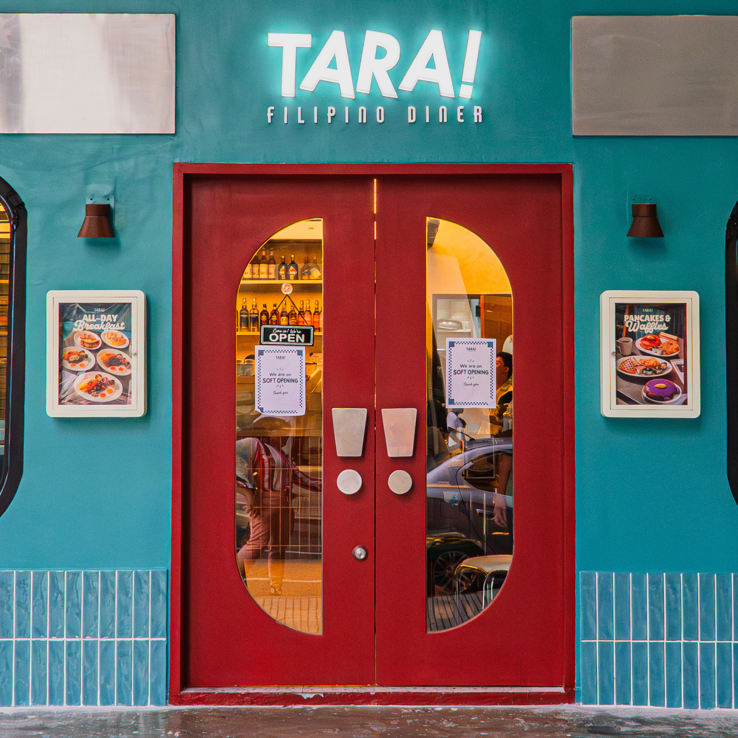 Teal Tara! Filipino diner storefront with bright red double doors, glowing sign, food photos, and window lettering advertising breakfast and coffee; inviting feel.